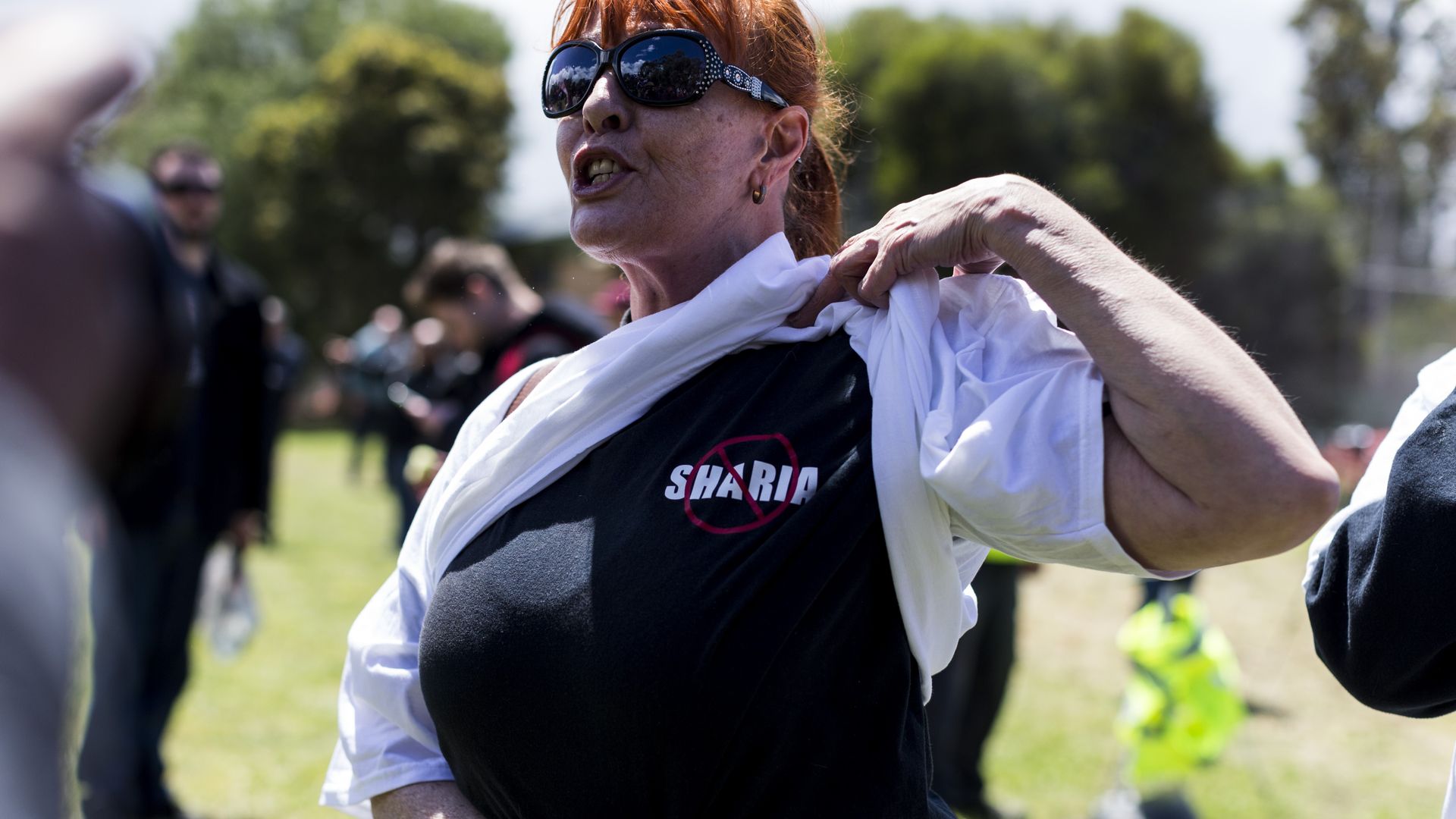 A woman reveals a shirt under her sweatshirt that has a red "no" symbol over the word Sharia.