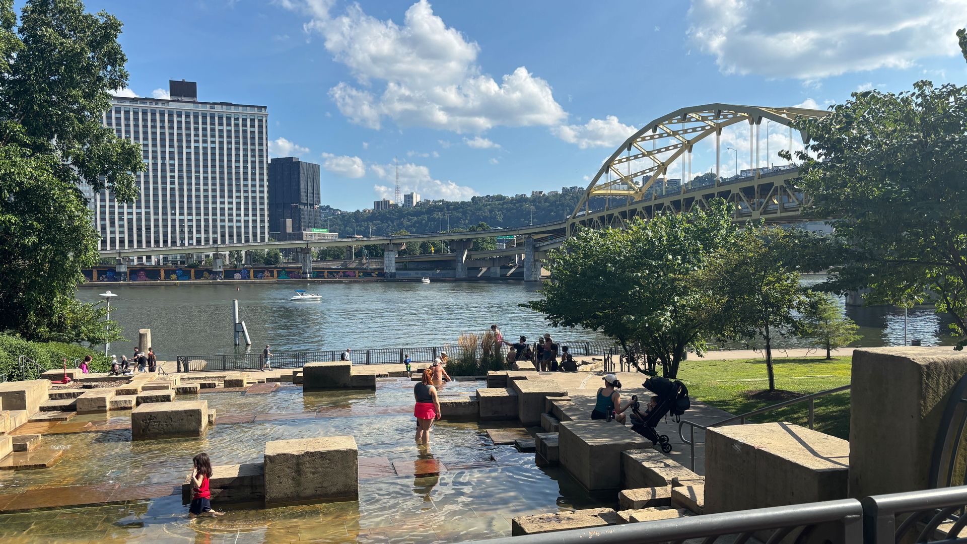 People enjoying a sunny day by a river with shallow water and concrete blocks, a yellow arch bridge, tall buildings, trees, and blue sky with clouds in the background.