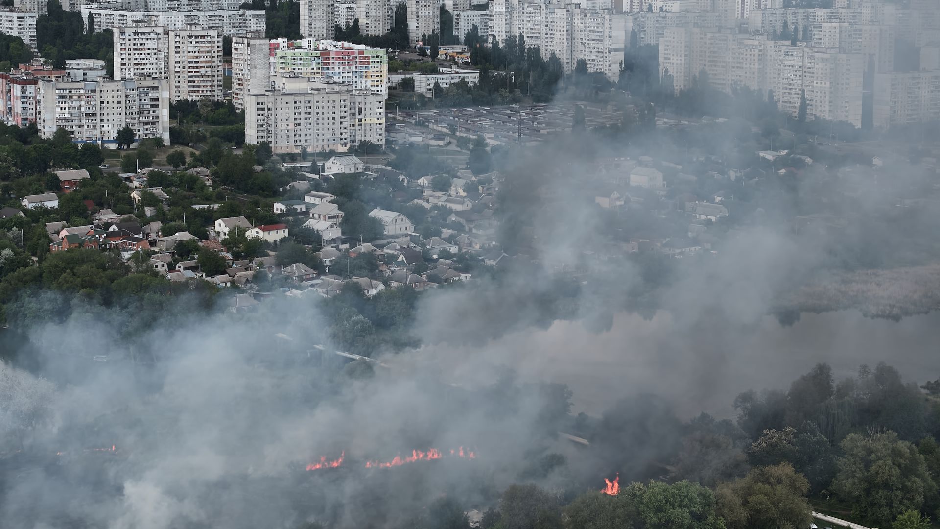 A birds-eye view photo of a city with tall buildings in the background, smaller houses in the foreground. Smoke covers the front of the image. Some trees are seen on fire. . 