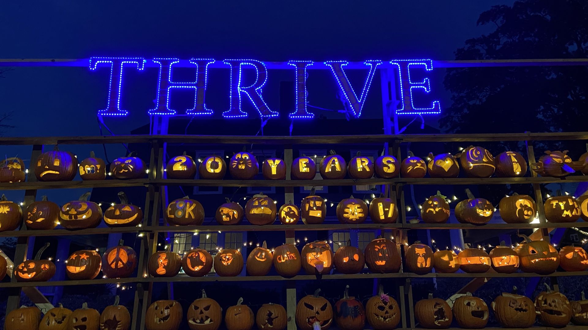 Rows of lit carved pumpkins at night with various designs and letters spelling "20 YEARS" beneath large blue lights forming the word "THRIVE".
