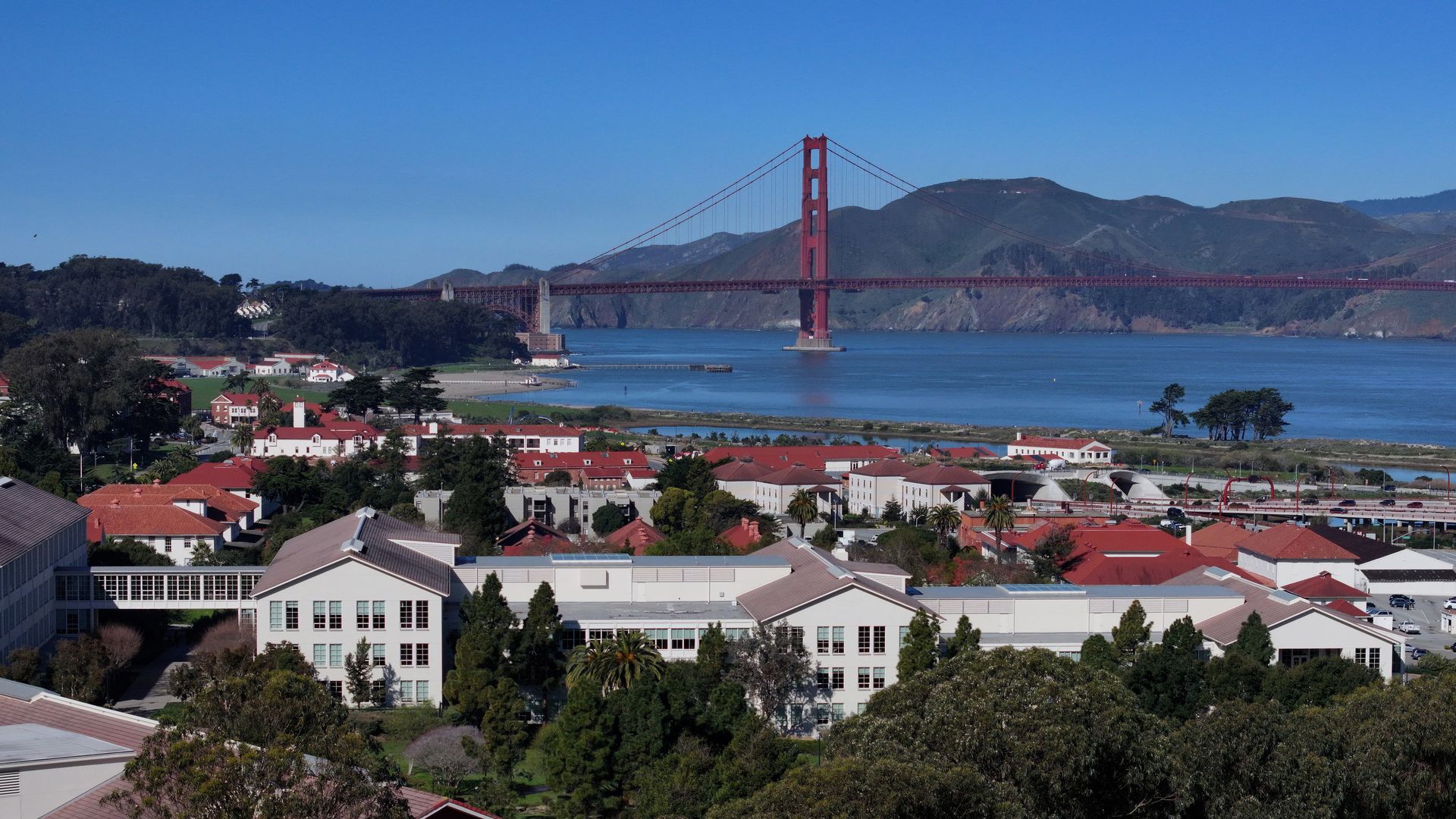 View of the Golden Gate Bridge in red, spanning blue bay with hills in the background; foreground shows a town with white buildings and red-tiled roofs, green trees.