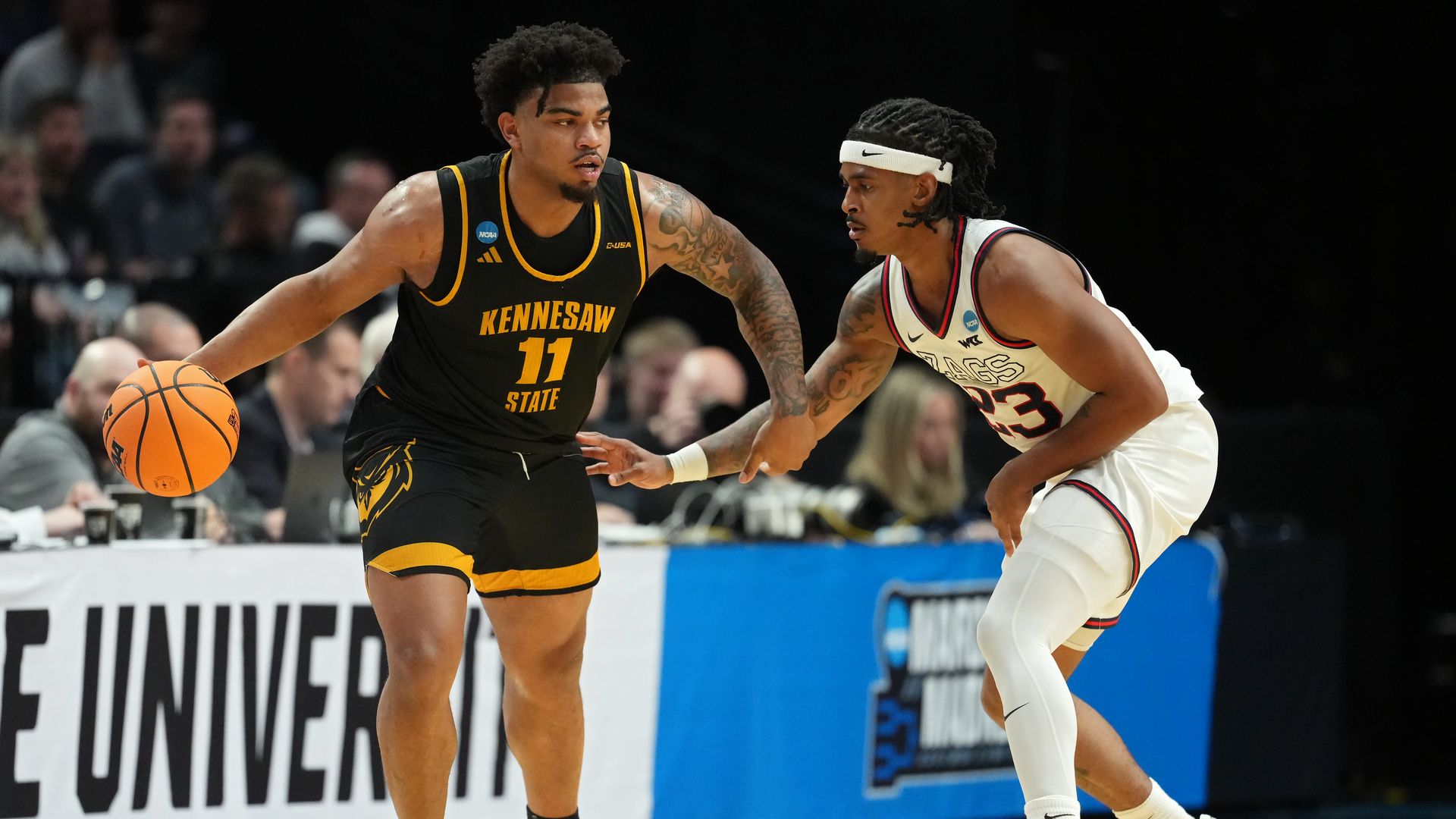 Two basketball players, one from Kennesaw State and one from Gonzaga University, are shown on the court, with the crowd in the background.