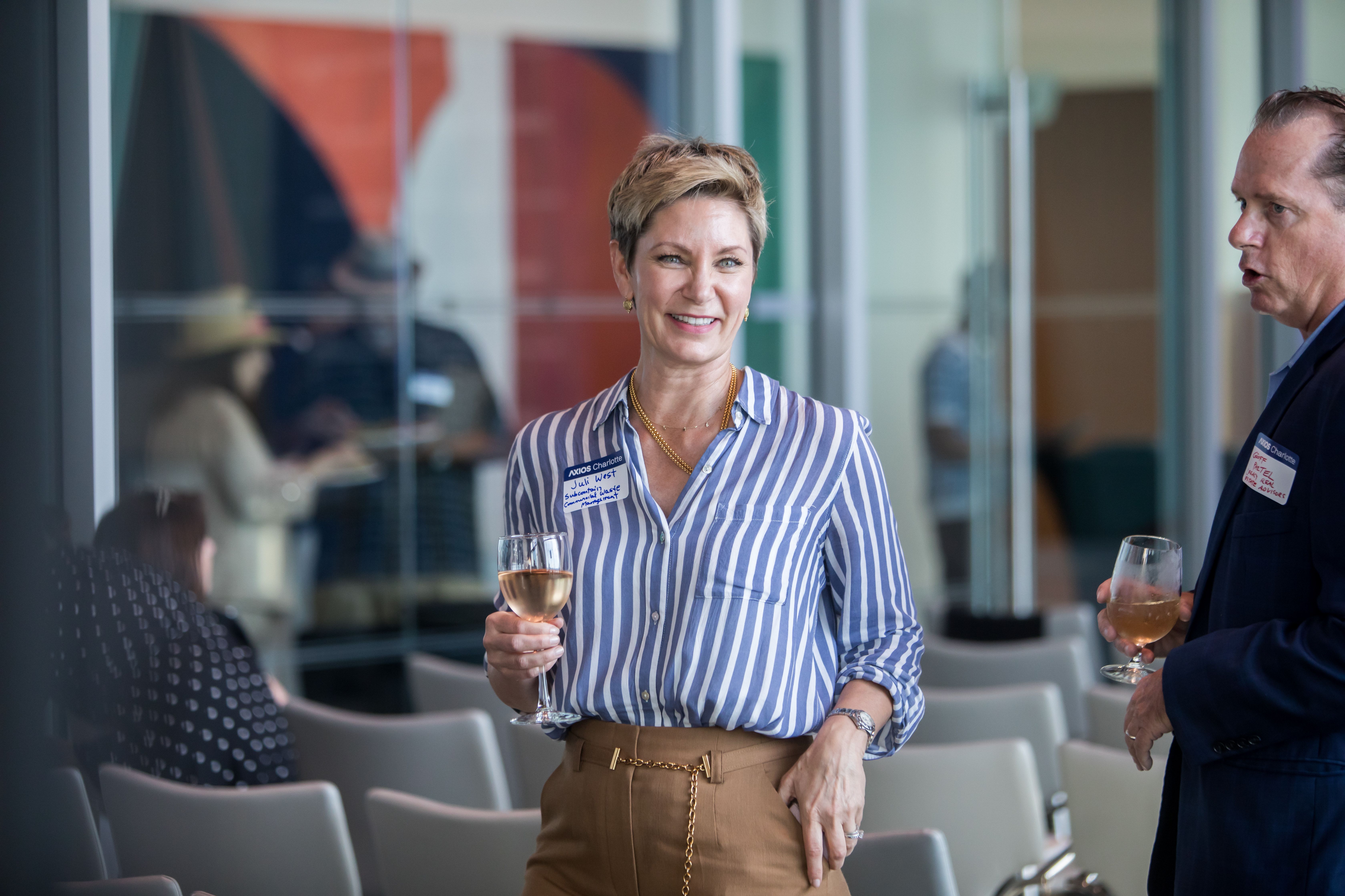 Smiling woman with short blond hair in a blue-and-white striped blouse holding a wine glass; a man in a dark suit speaks in the background inside a glass-walled conference space.