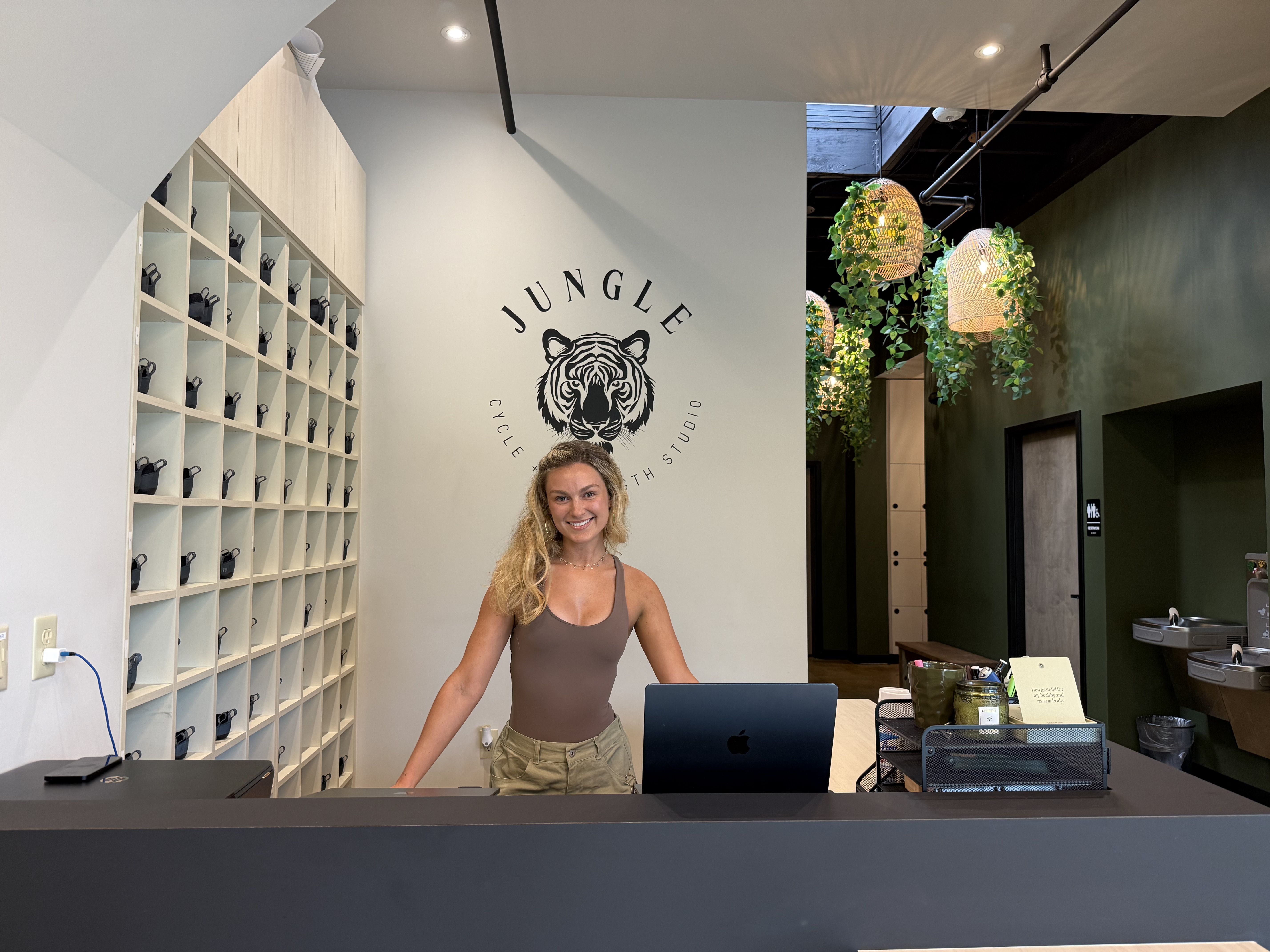 Reception at Jungle Health Studio: smiling blonde staff at a dark counter, wall logo with tiger and curved text, cubby shelves with small bags, woven pendant lights, and trailing green plants.
