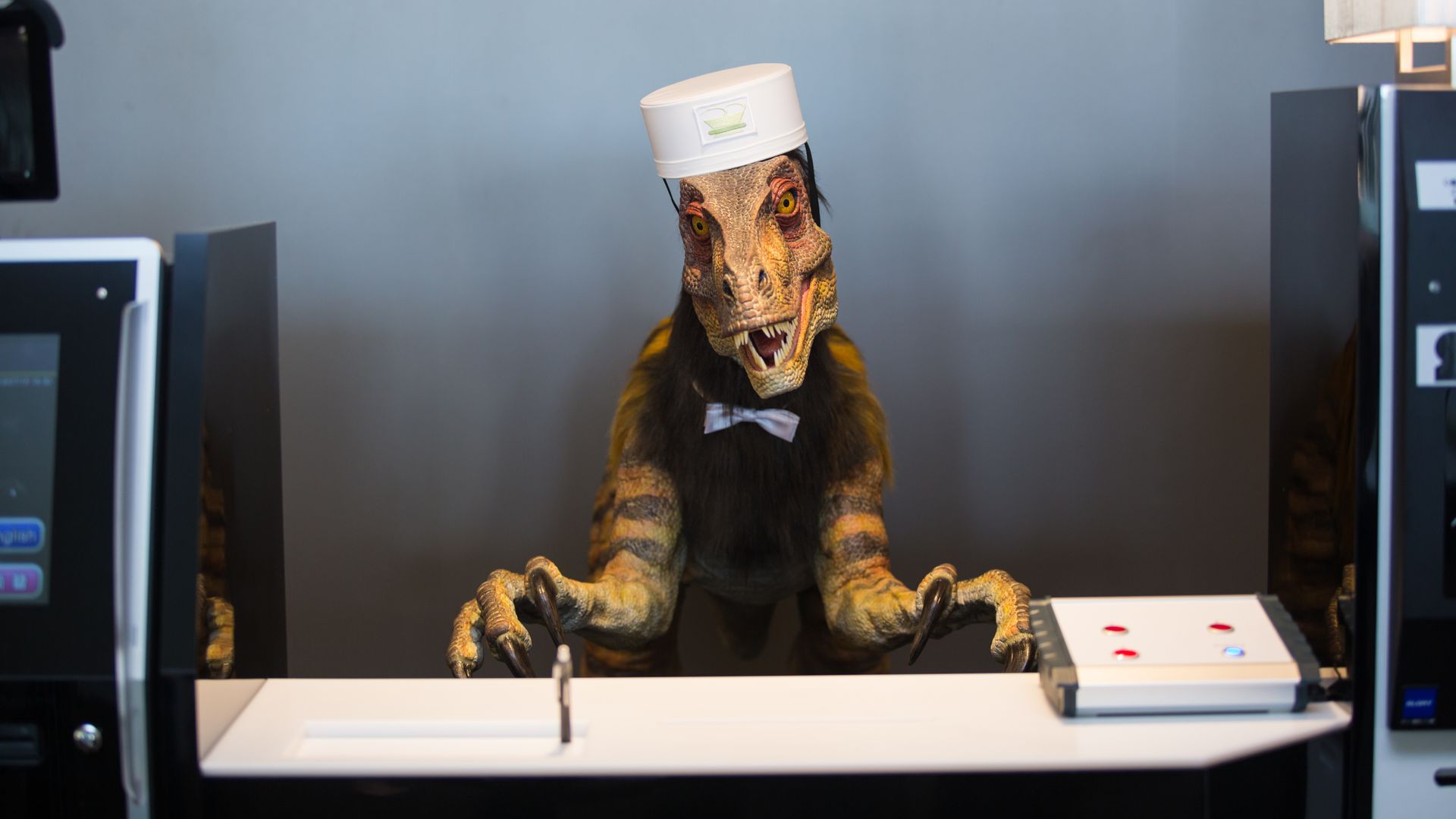 Photo of a robotic dinosaur wearing a cap and uniform behind a concierge desk