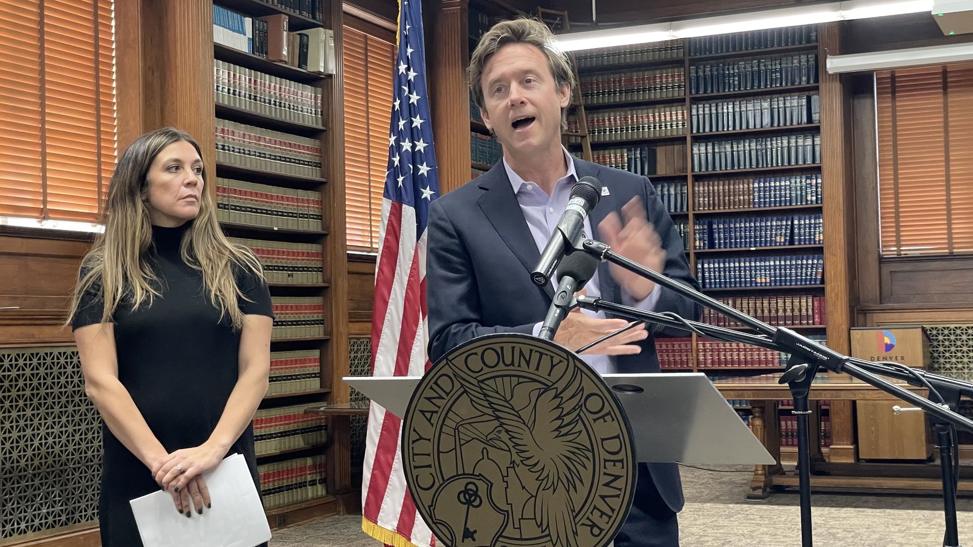 A man speaks in front of a lectern while moving his hands, while a woman watches next to him.