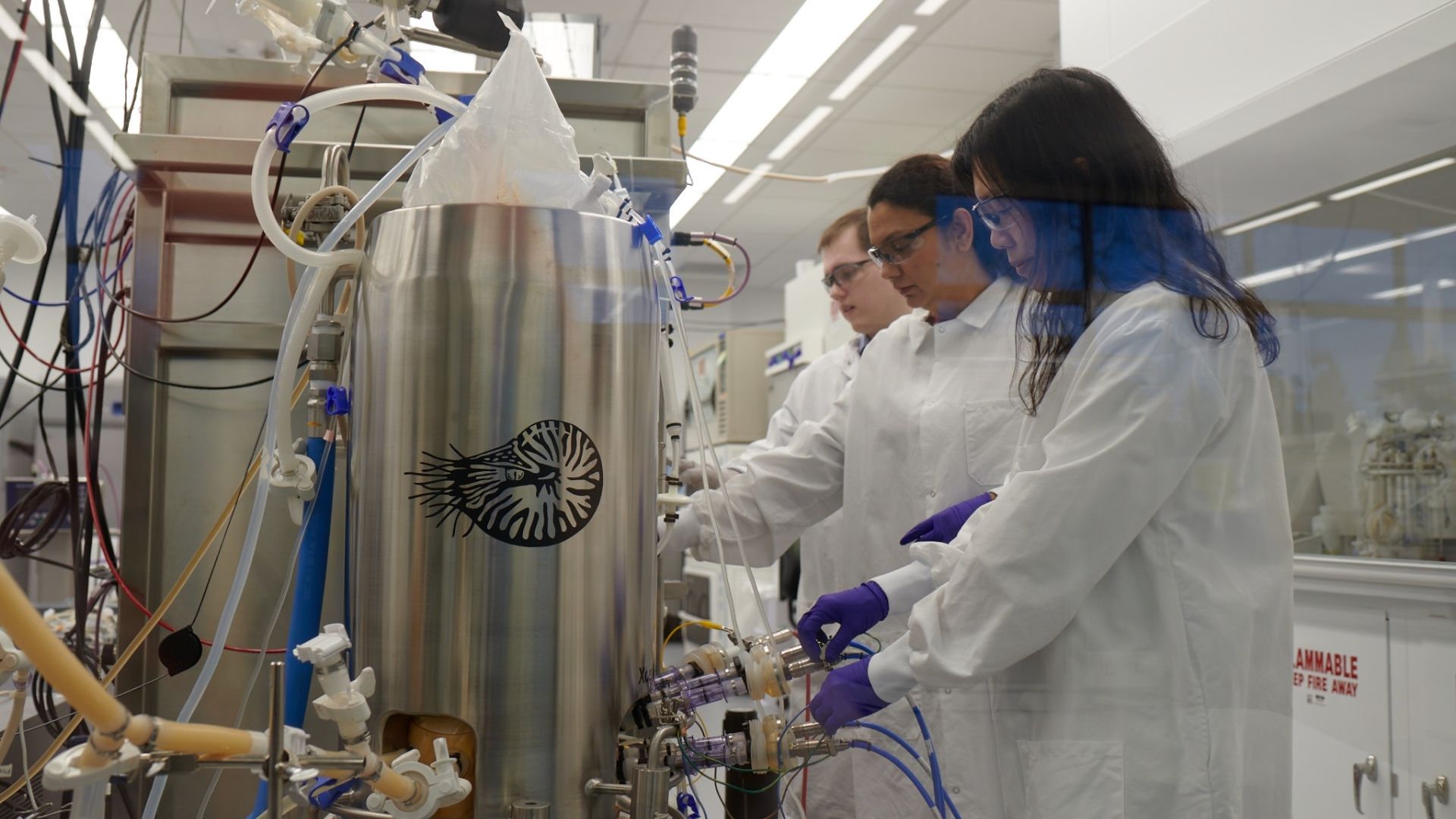 Three scientists in white lab coats and purple gloves work at a stainless steel reactor in a modern lab, surrounded by tubing and glassware, with safety gear and a spiral shell logo on the vessel.