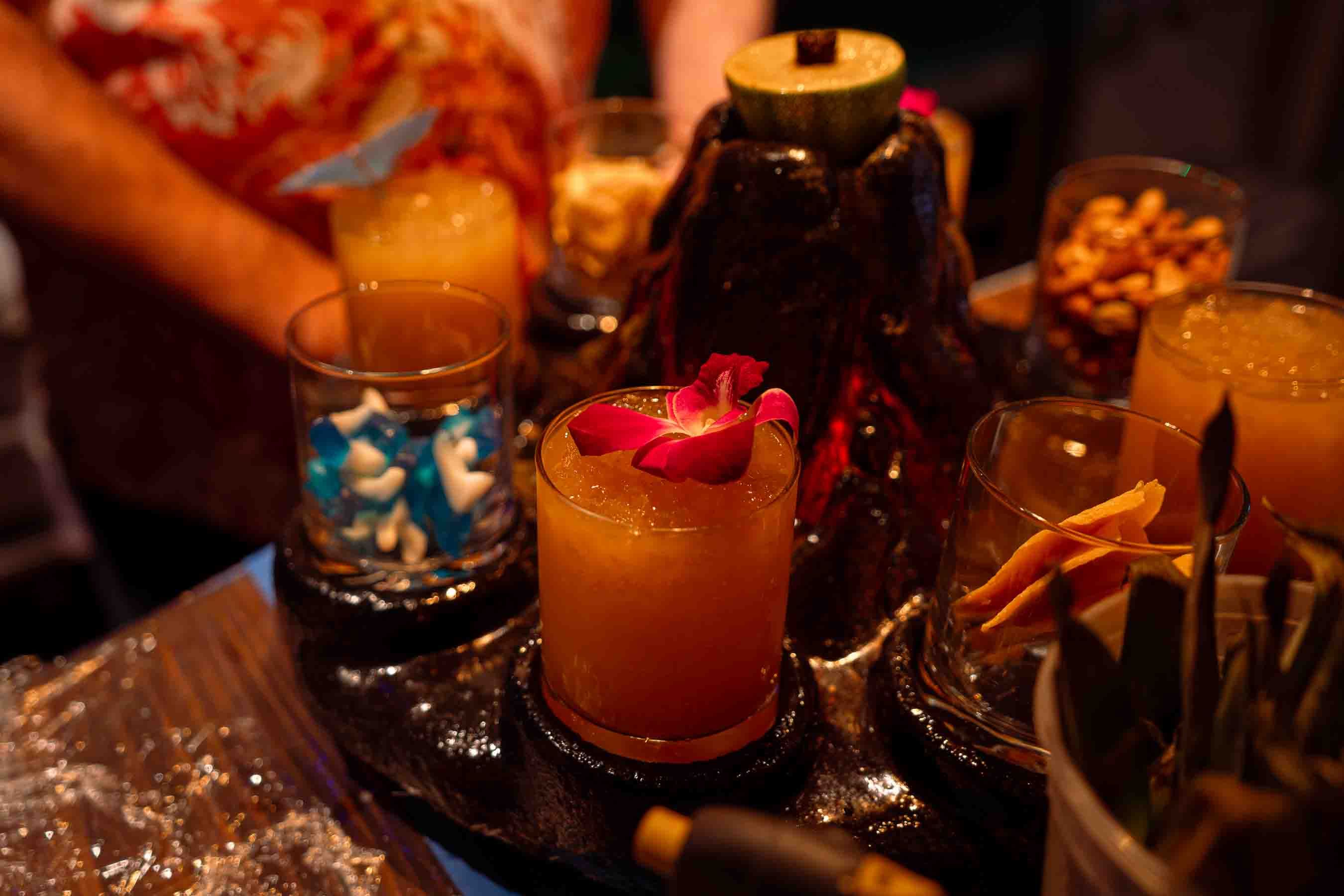 Dimly lit party bar scene with an orange cocktail garnished by a pink orchid in a lowball glass in the foreground. A dark bottle and bowls of nuts surround the drinks.