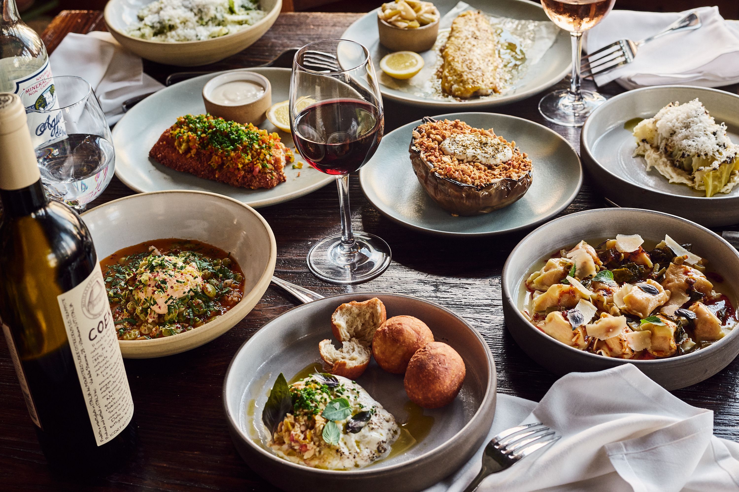 A close-up photo of a brown wooden table filled with a variety of dishes, a bottle of wine and a glass of wine.