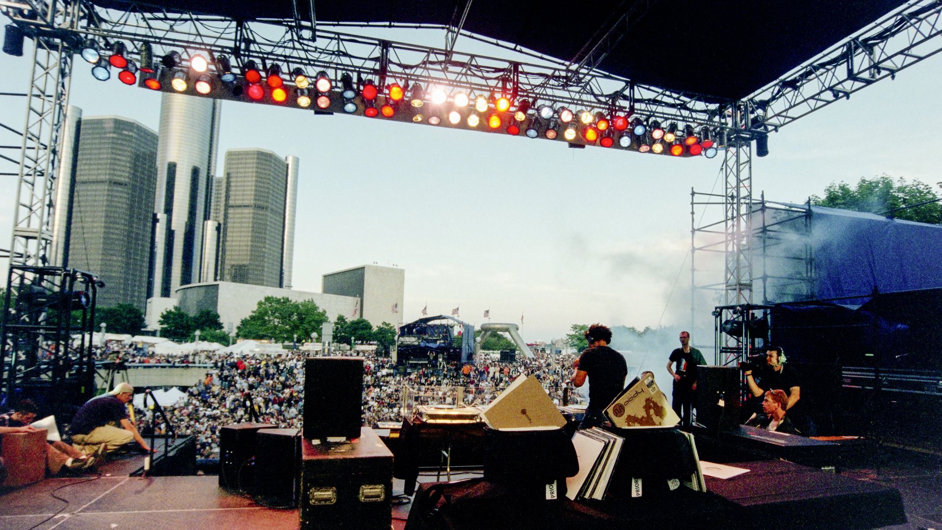 A techno artist faces a huge crowd with the RenCen visible behind.