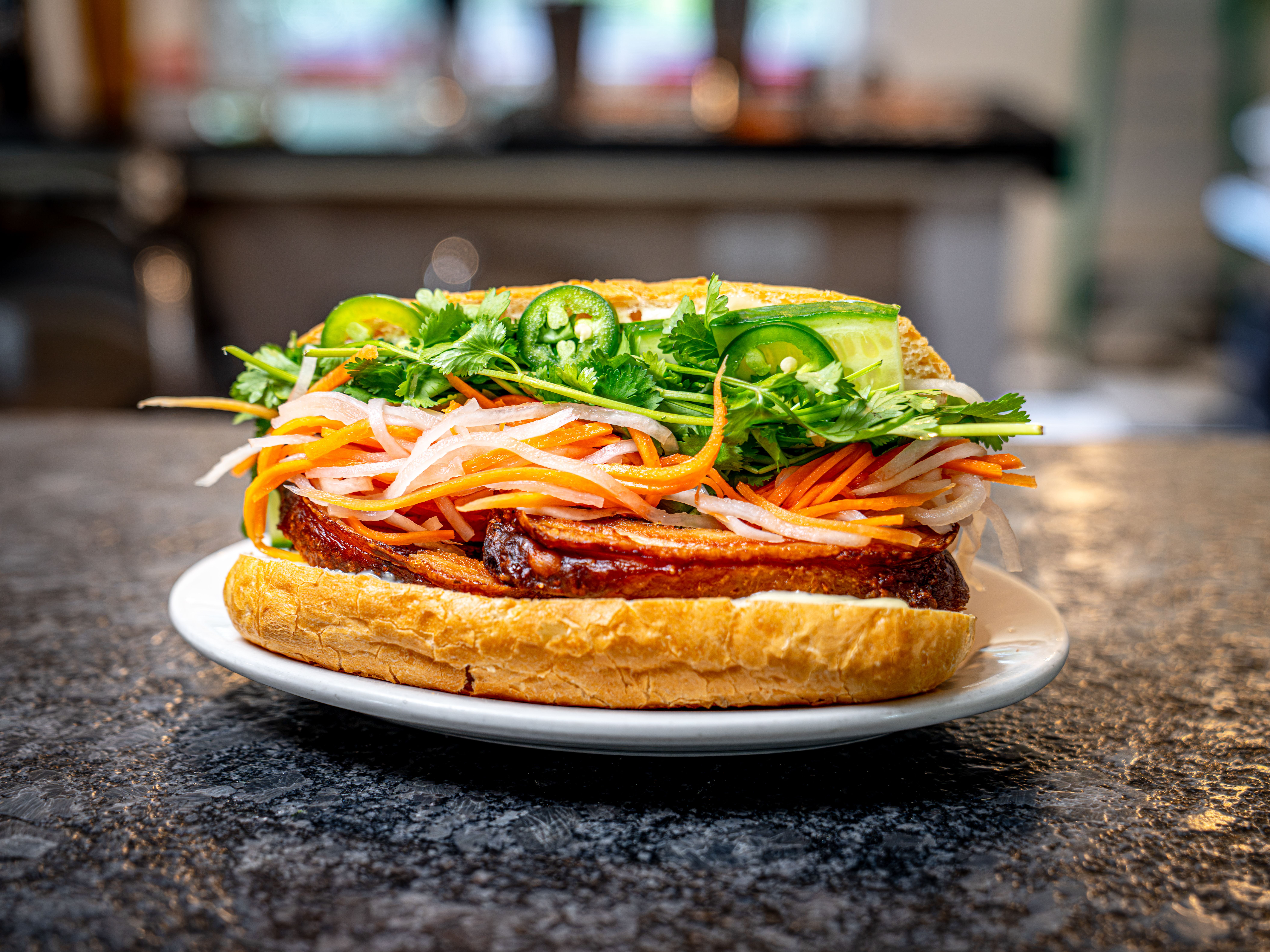Close-up of a toasted baguette sandwich piled with shredded carrot and daikon, cilantro, cucumber, and jalapeño slices; fresh greens on a white plate.