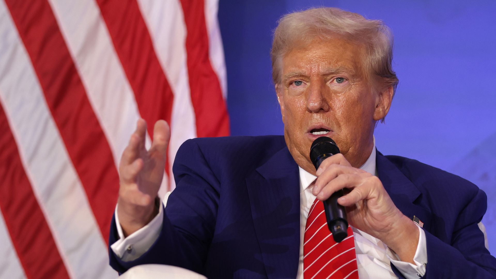 Donald Trump speaks holding a microphone with part of an American flag visible behind him