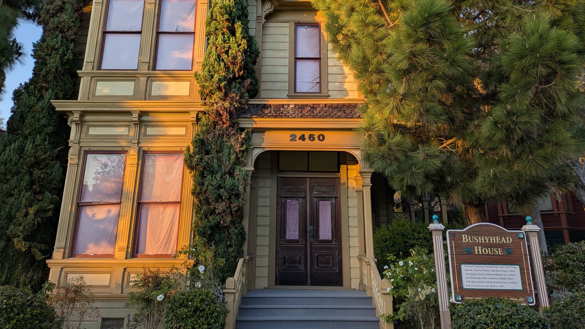 Two-story Victorian-style yellow house with brown trim, tall bay windows, and a dark wooden front door. Address 2460 above the entry. Surrounded by tall evergreens; a sign reads "Bushyhead House".