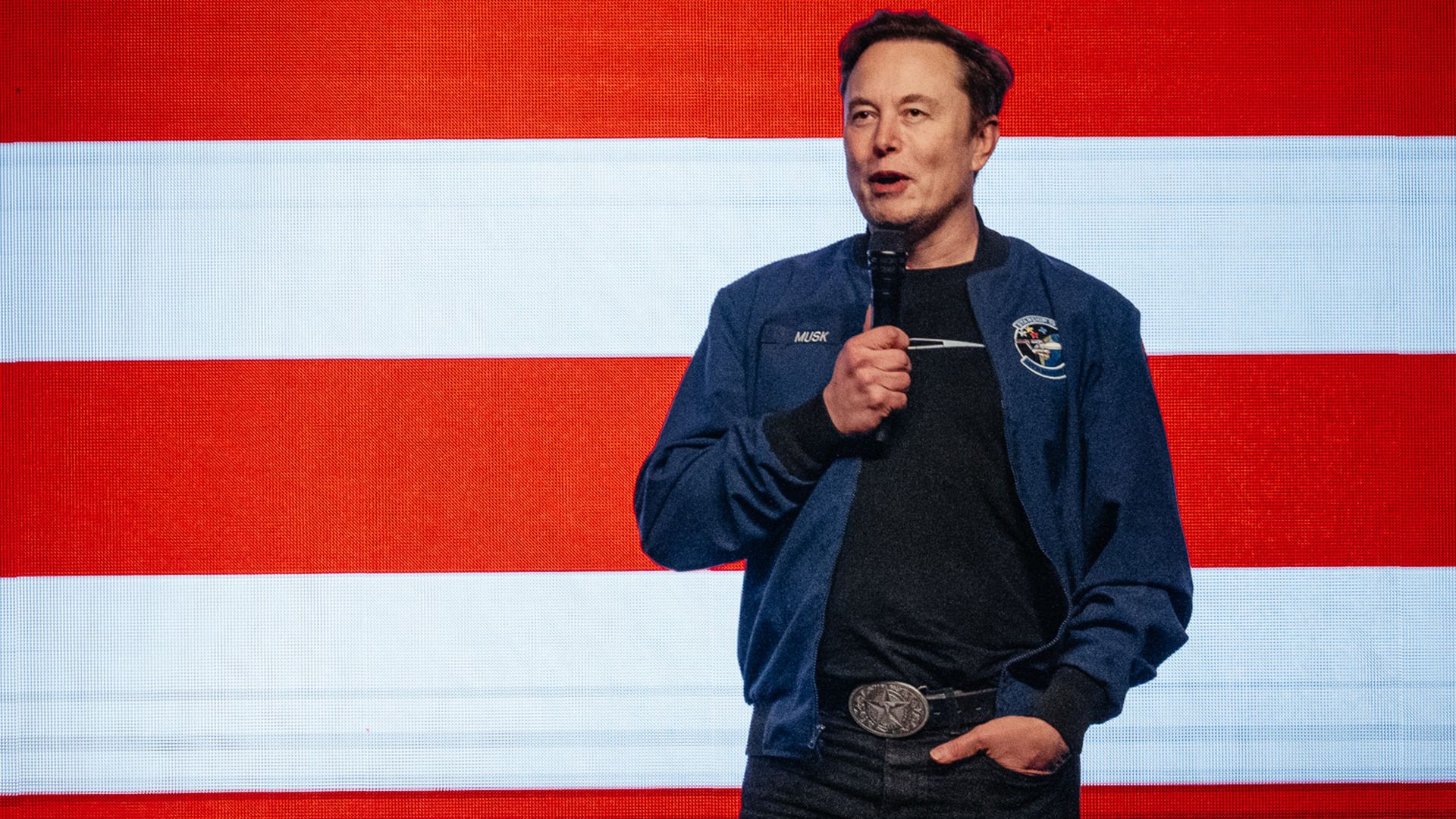 A person in a blue jacket speaks with a microphone in front of a screen with the American flag on it
