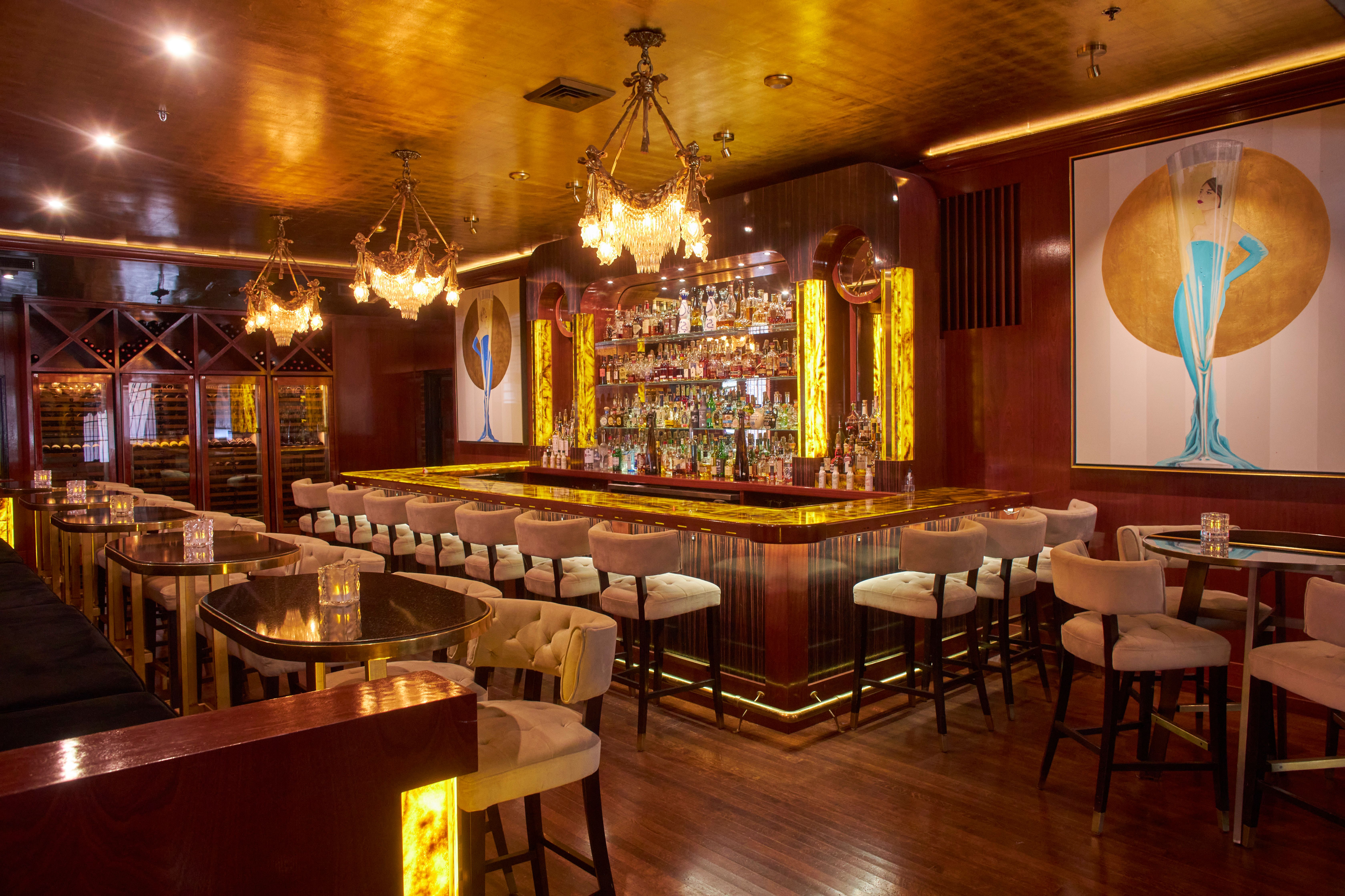 Luxurious bar with warm amber lighting, crystal chandeliers, gold ceiling, bottle shelves, a long gold-trimmed bar, beige stools, and a framed illustration of a blue-dressed woman in a gold circle.