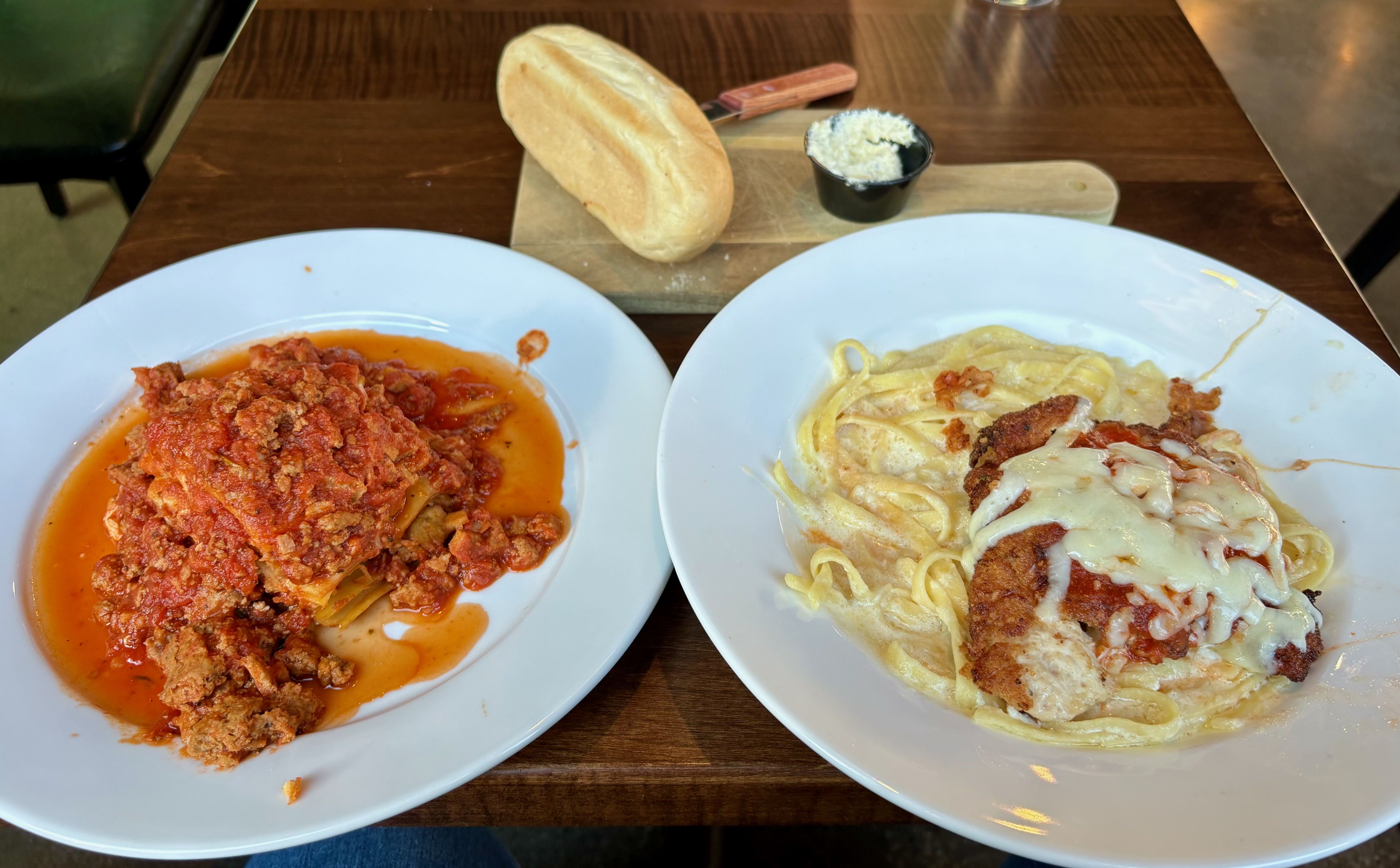 Two white plates, with lasagne and fettucine alfredo with chicken parmigiana, and a loaf of bread on a cutting board