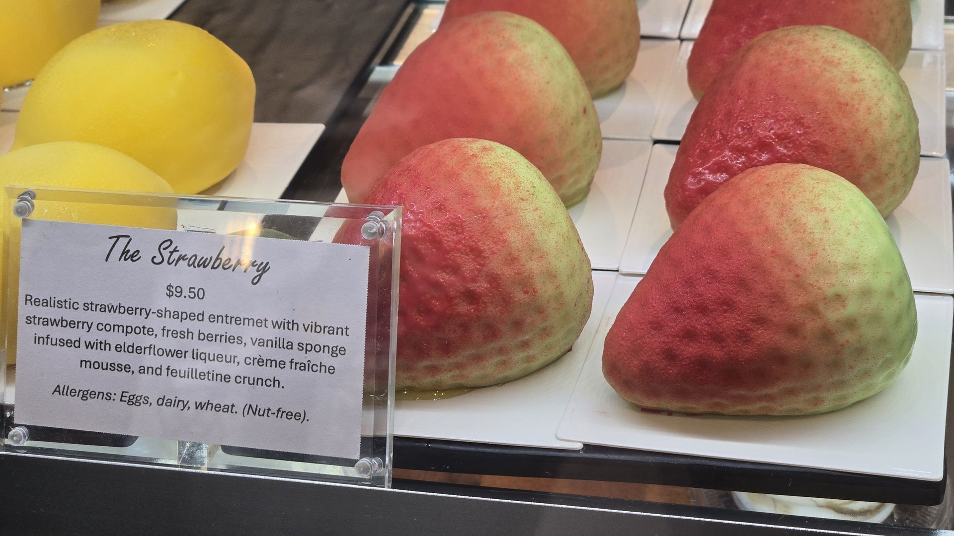 Close-up of a fruit display with red and green mangoes on white shelves; a glass sign reads "The Strawberry" and $9.50, with a detailed description of the dessert.