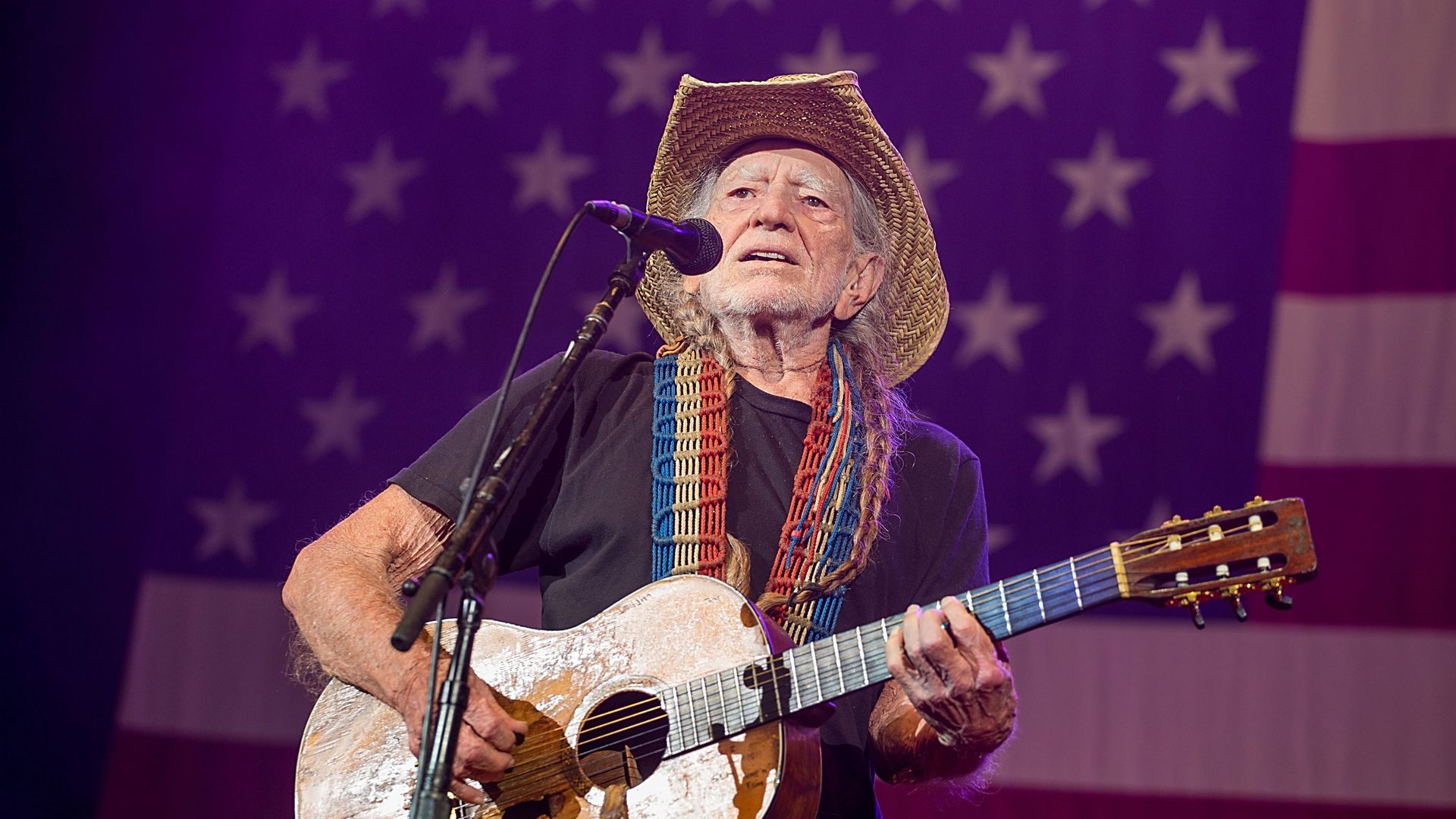 Older man in a straw hat plays a weathered acoustic guitar on a purple-lit stage, with a microphone in front and a blurred United States flag backdrop.