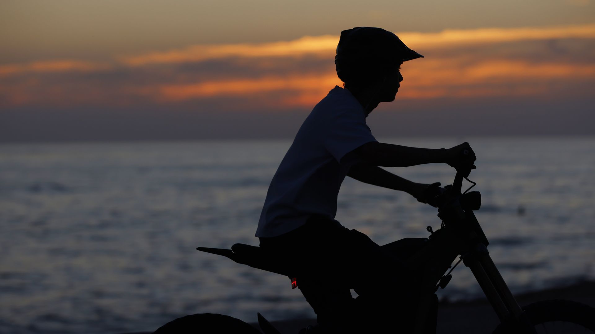 A silhouette of a teenage boy wearing a helmet riding an e-bike along the coast with a sunset in the background.