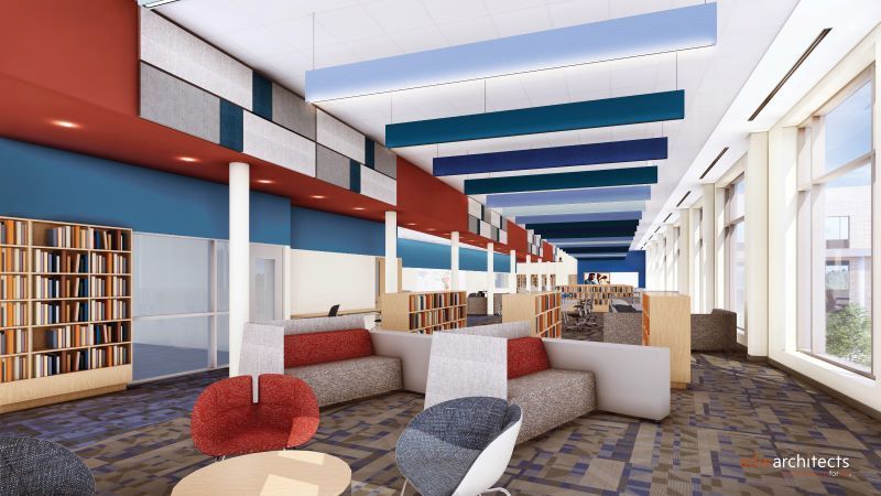 Modern library interior with red and blue accent walls, large windows letting in natural light, bookshelves, gray and red cushioned seating, and suspended blue acoustic panels on the ceiling.