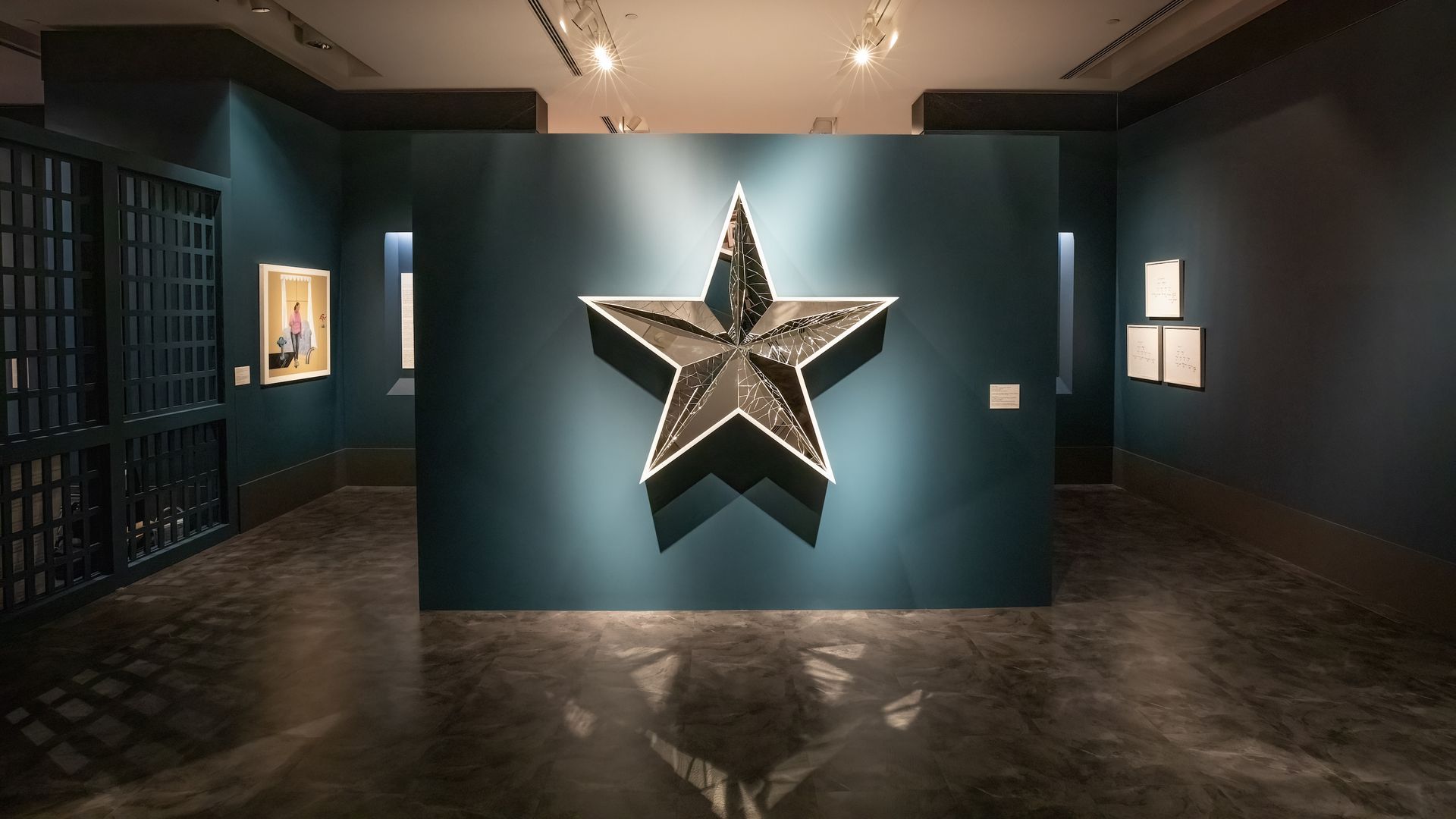 A silver star-shaped artwork hangs on a teal blue wall in a gallery.