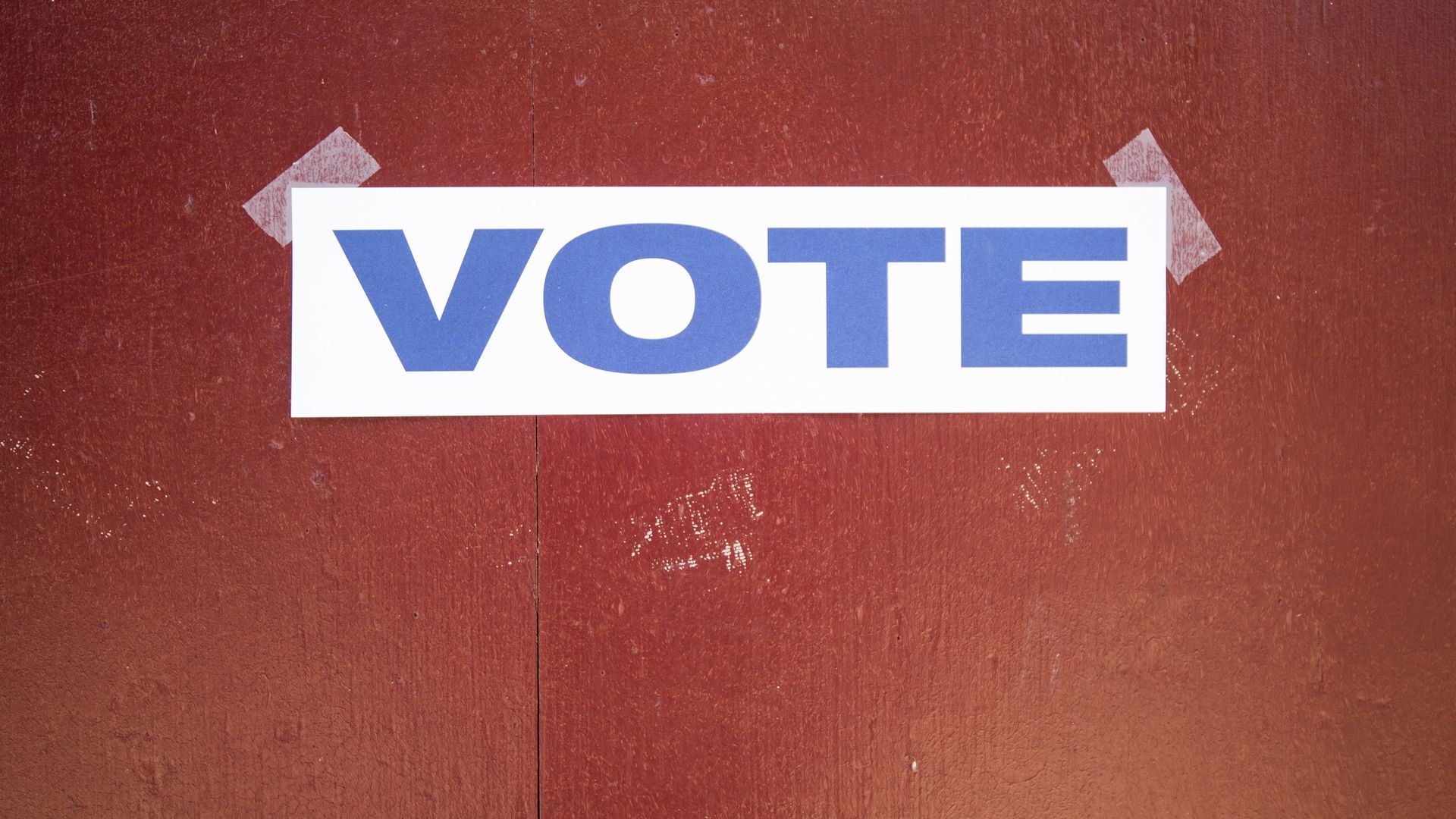 Sign that says vote in all caps in blue on white paper taped to red wood door.