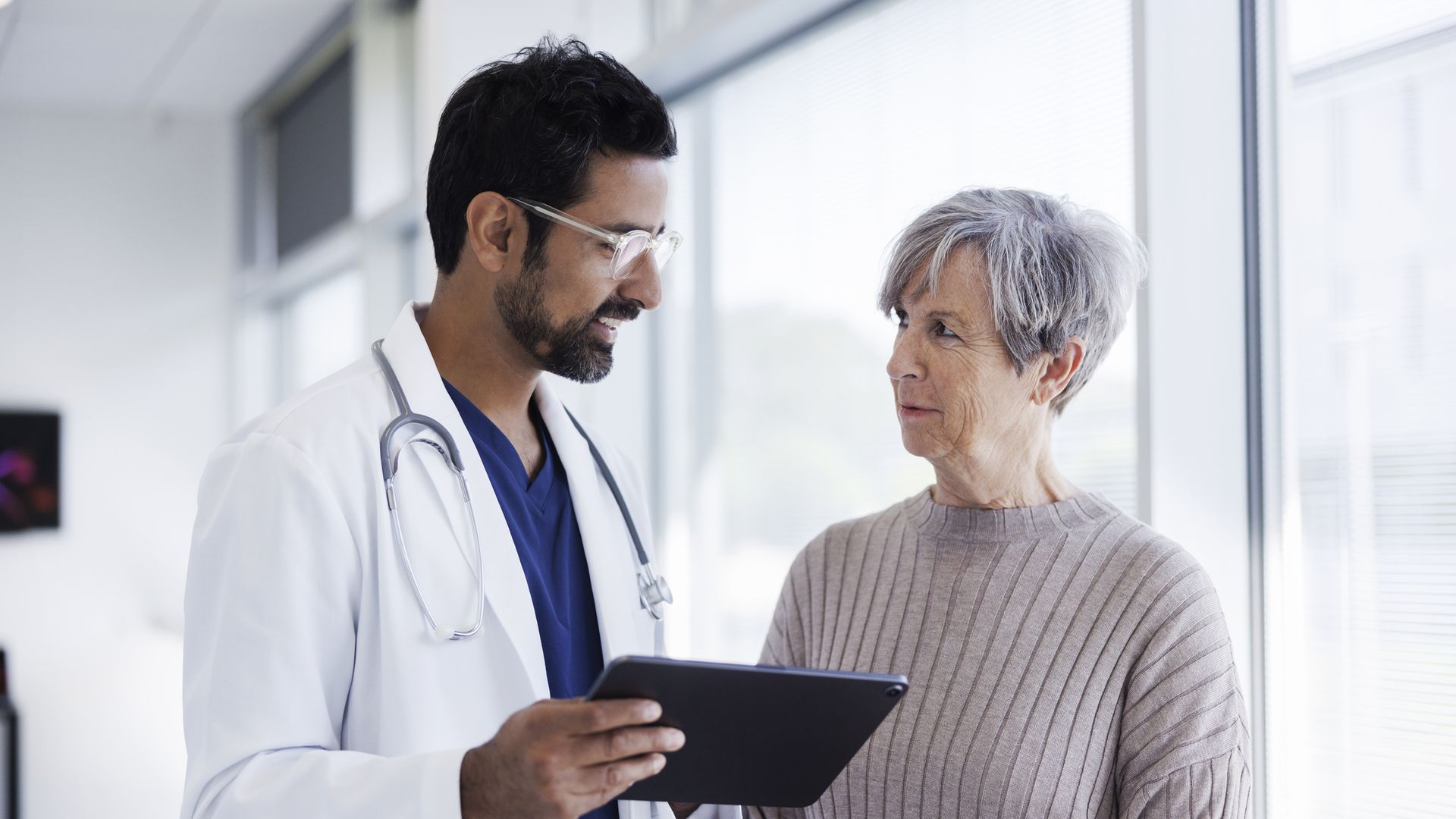A male doctor with stethoscope and glasses shows a tablet to an elderly woman with short gray hair wearing a light sweater, as they talk beside bright clinic windows.
