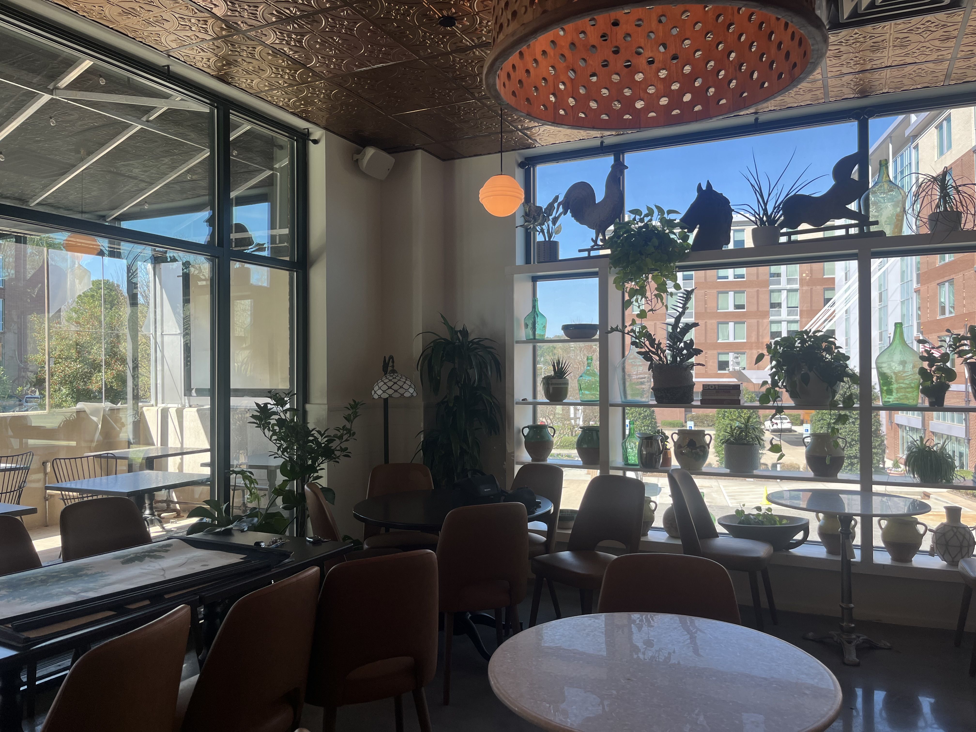 Bright cafe interior with large windows, brown chairs, and white round tables. Window shelves hold green glass bottles and potted plants; outdoor patio visible; copper ceiling adds warmth.