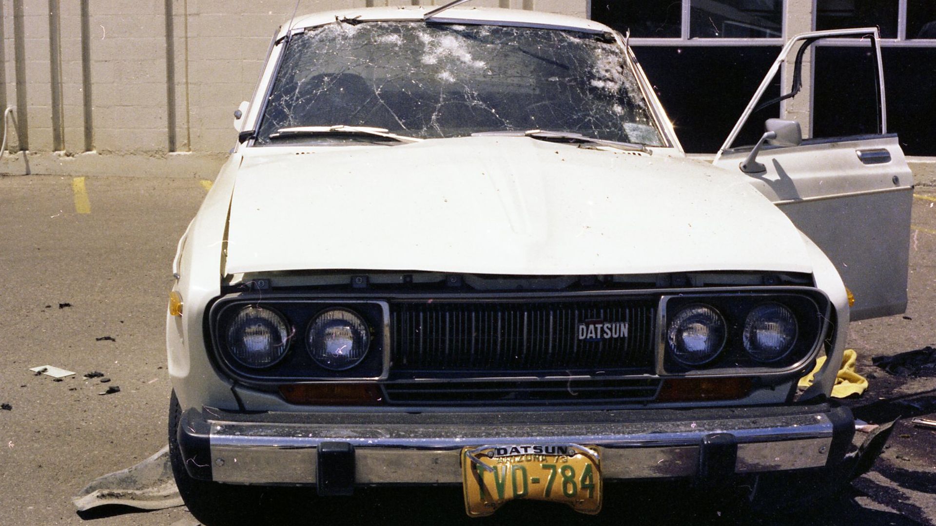 Front view of a white Datsun with a cracked windshield and dented hood; driver's door open, yellow license plate VD-784, debris on the ground in a parking lot beside a building.