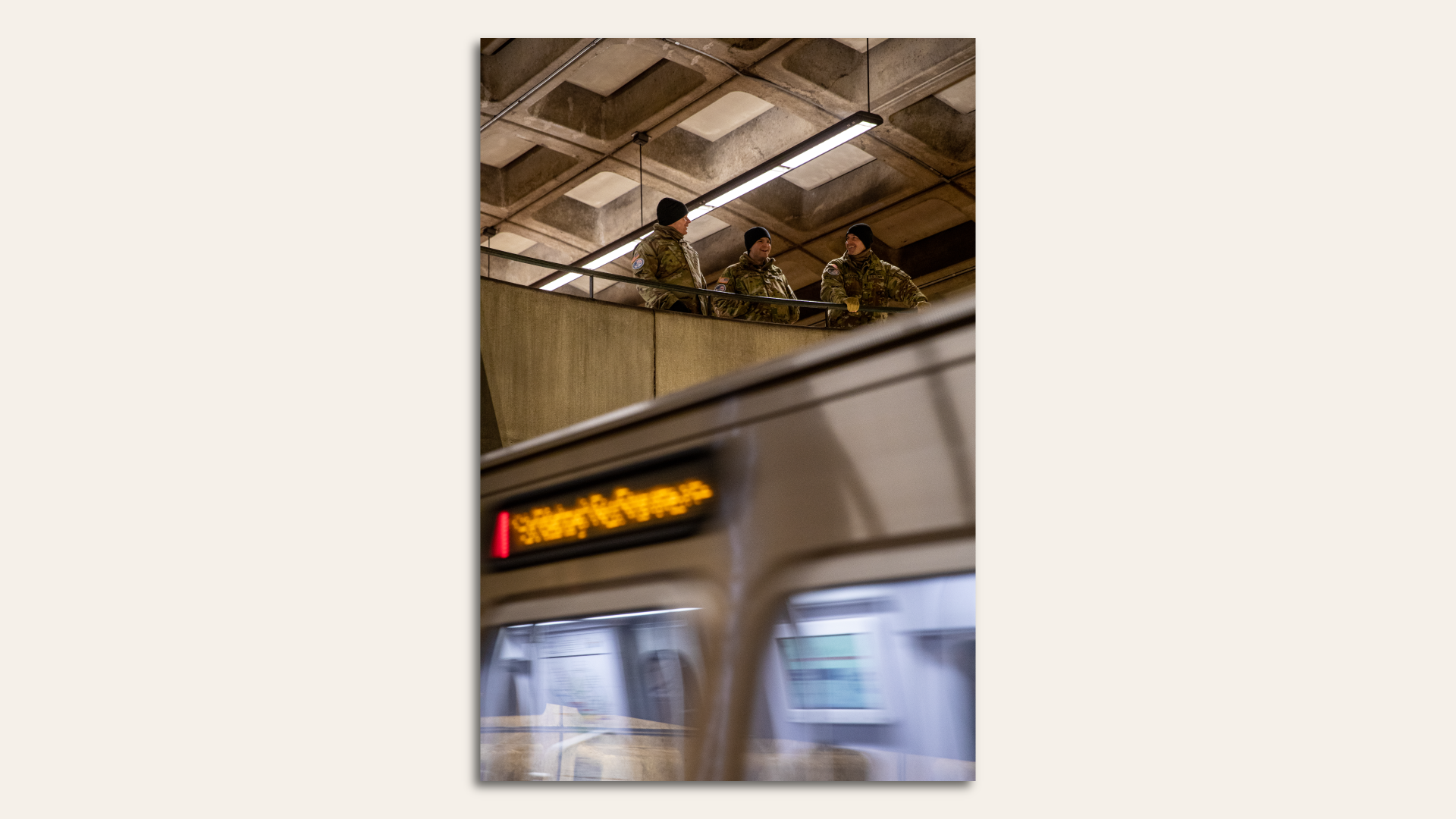 Three men in military uniforms and beanies stand on a second floor of a subway station as a train pulls in.