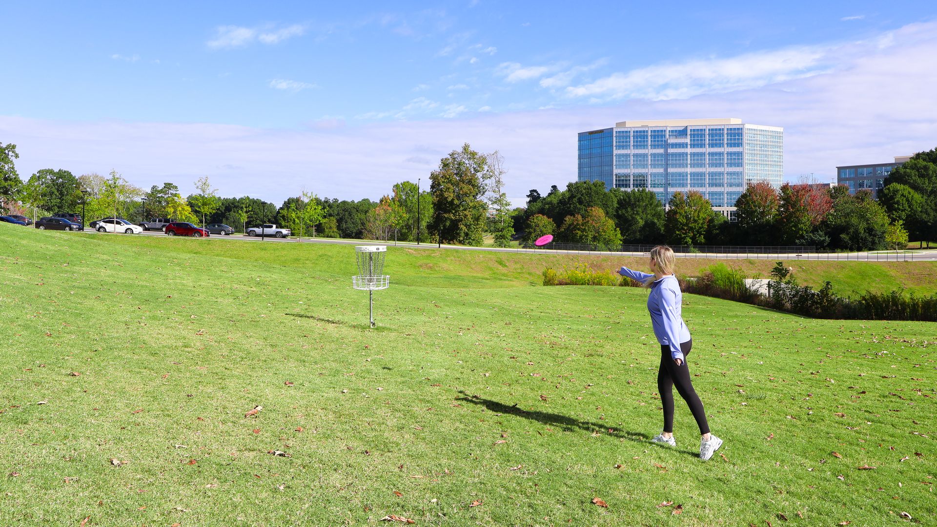 Person in blue shirt and black leggings throwing a pink disc towards a disc golf basket on a grassy field with trees and a glass building in the background under a blue sky.
