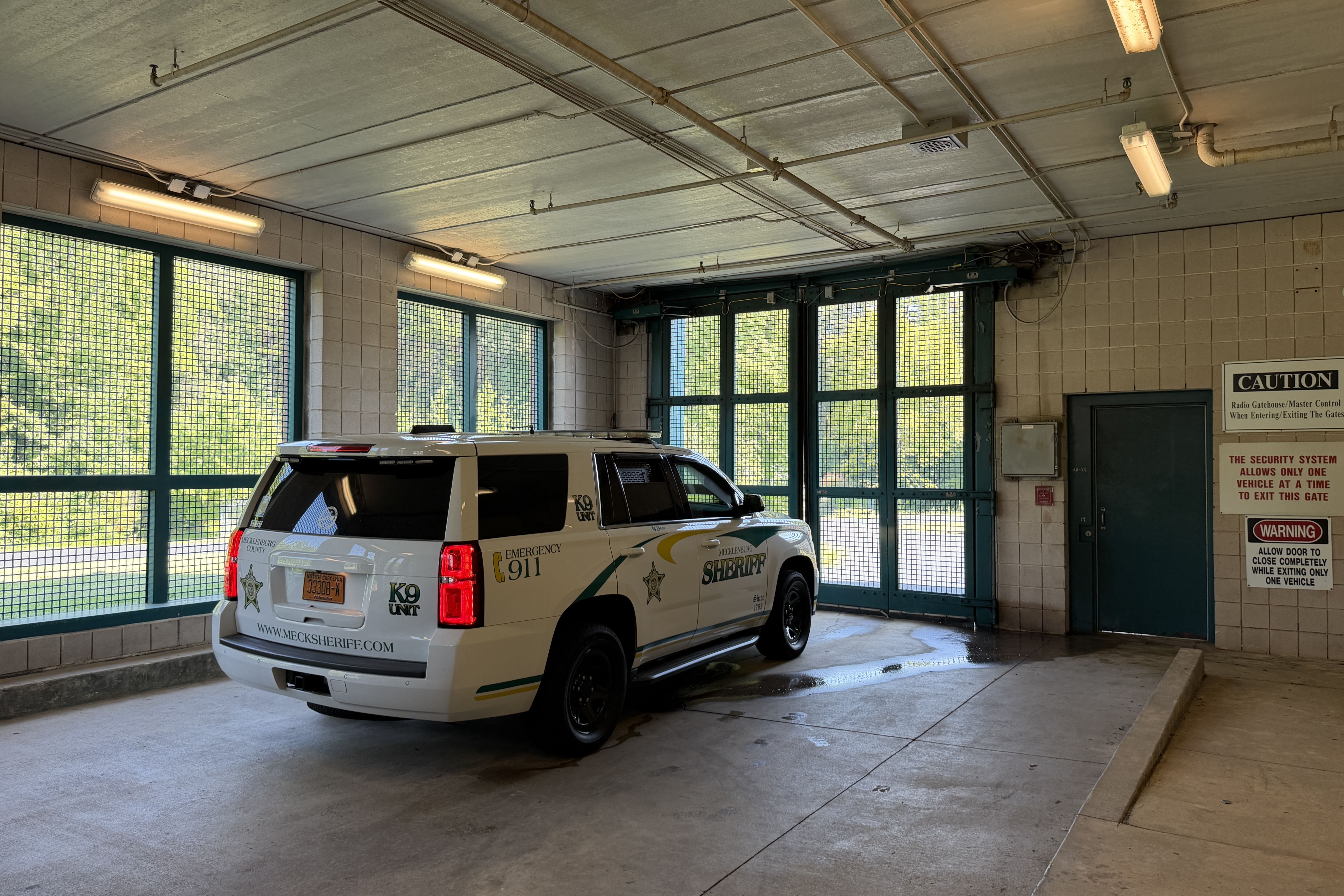 Interior of a police garage: a Mecklenburg County Sheriff SUV parked near teal-framed windows; exposed ceiling pipes, fluorescent lights, tiled walls, and caution signs by a large door.