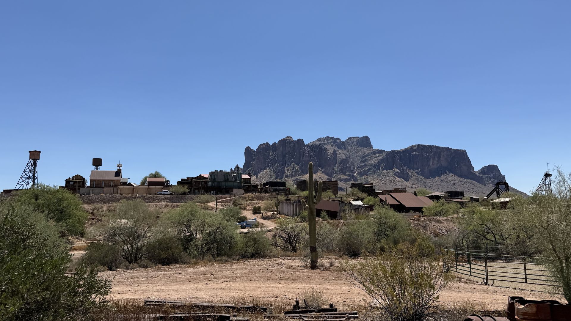 Desert frontier town with weathered wooden buildings and rusted mining towers, set against a rugged mesa under a bright blue sky; foreground shows dry scrub and a cactus.