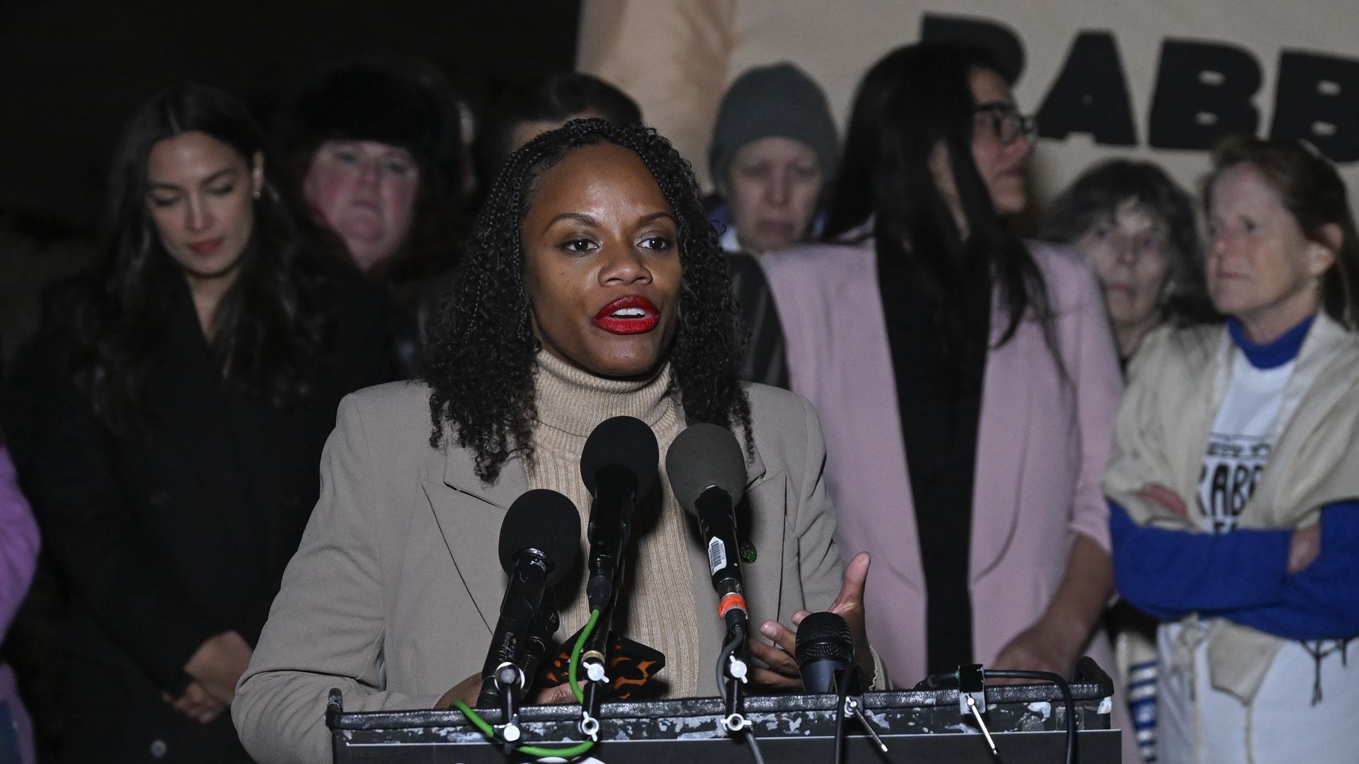 Rep. Summer Lee, wearing a tan blazer and tan turtleneck, speaking into a microphone surrounded by colleagues and supporters.