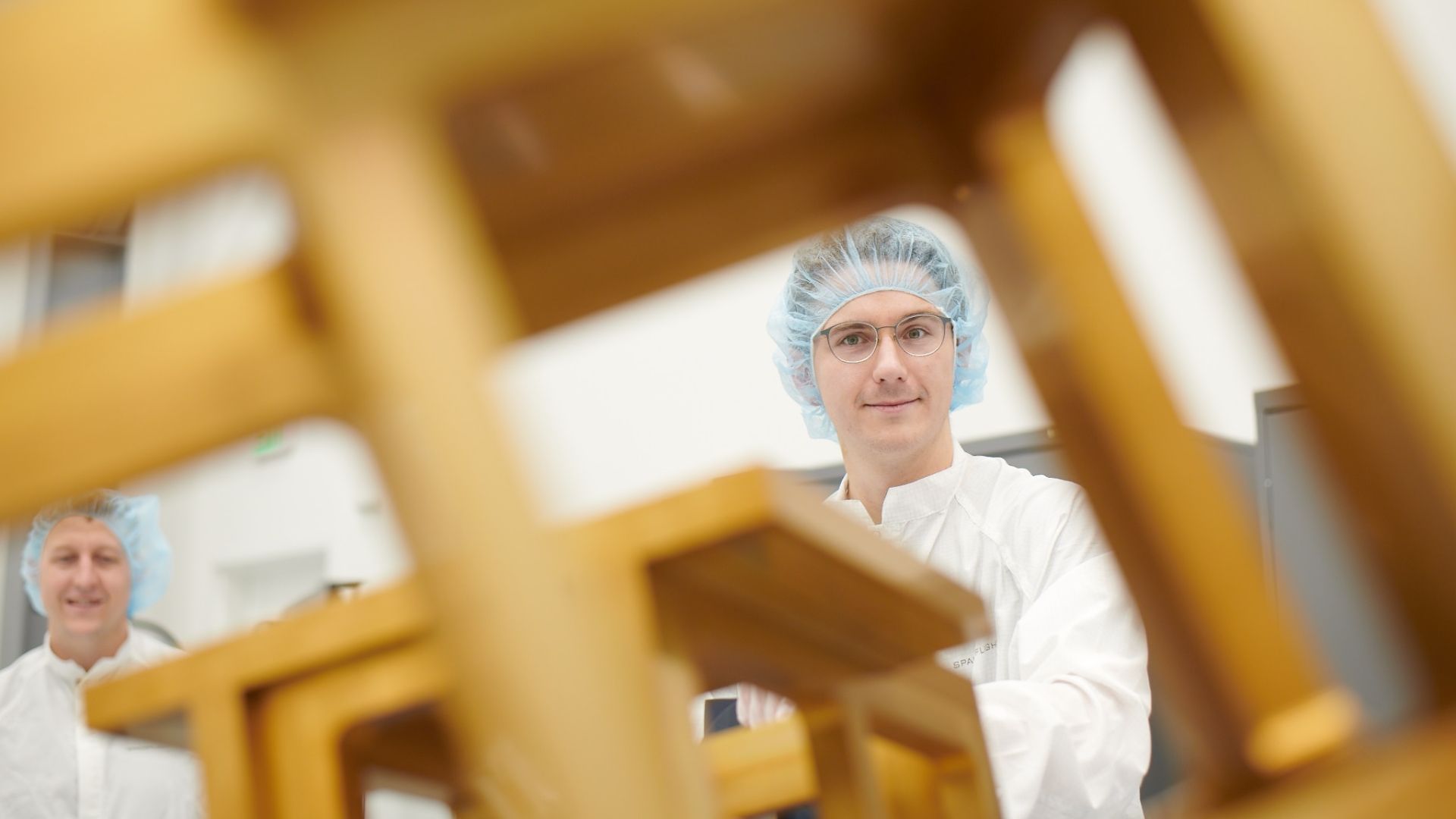 Man in a white lab coat and blue hairnet with glasses smiles at the camera in a lab-like setting; another worker in a hairnet is blurred in the background, with wooden beams framing the scene.
