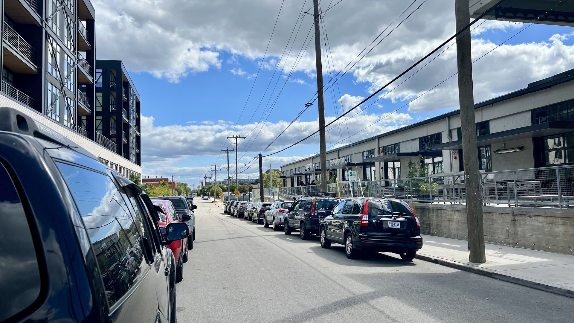 Urban street scene with cars parked on both sides, modern black buildings on the left, white industrial-style buildings on the right, under a partly cloudy blue sky.