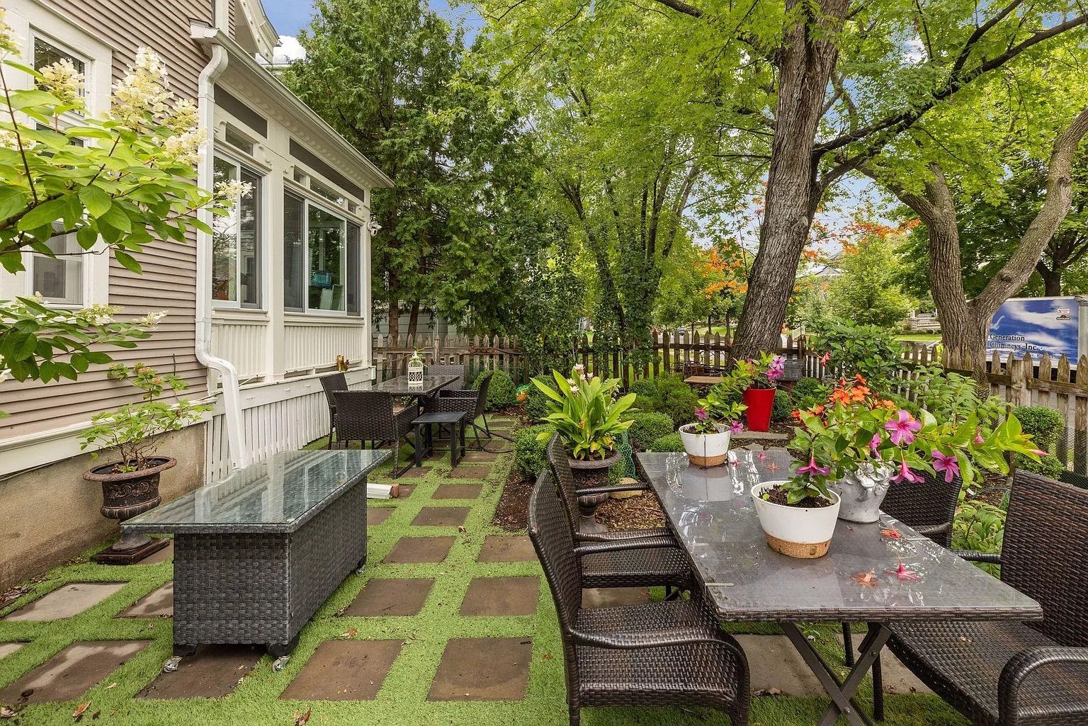 Outdoor patio with dark wicker chairs and two glass-top tables adorned with potted plants and pink flowers, surrounded by lush green trees and a wooden fence near a beige house.