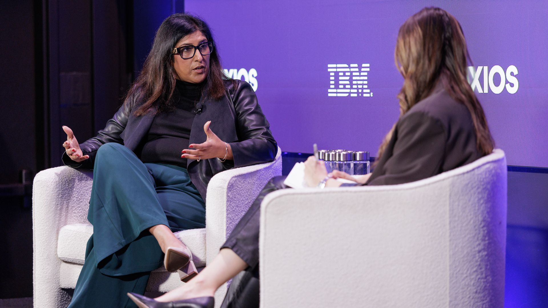 Two women sit on white chairs in a panel discussion on a stage with a purple IBM backdrop; the left speaker wears glasses, a black top and leather jacket, gesturing as the other woman faces away.