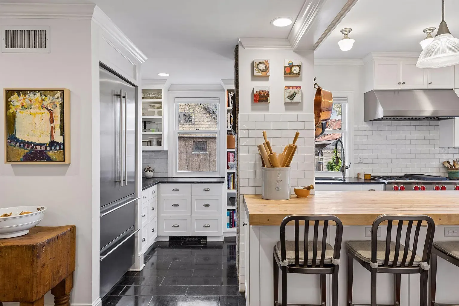 Bright white kitchen with a wooden island, two metal stools, and a large stainless refrigerator. White subway tile, pendant lights, and a can of rolling pins on the island.