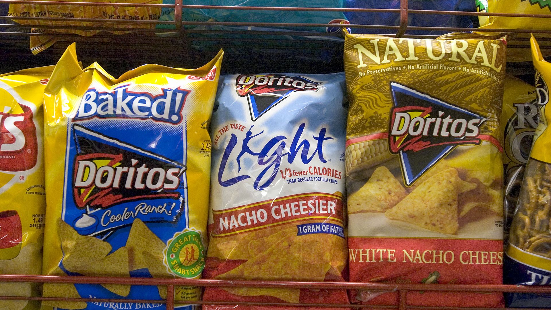 Row of large chip bags on a store shelf: left shows Baked! Doritos Cooler Ranch in yellow and blue, center Doritos Light Nacho Cheese in blue/white, right Doritos White Nacho Cheese in gold.