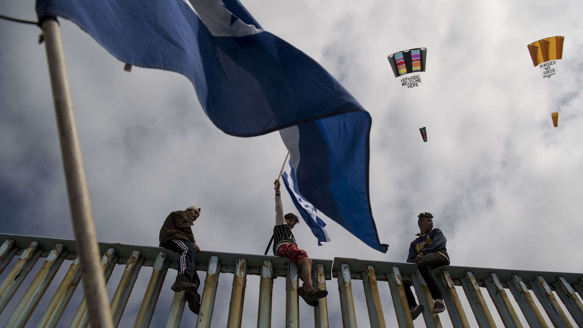 Three immigrants from Central America sit on top of the border wall with a flag flying in the foreground. 