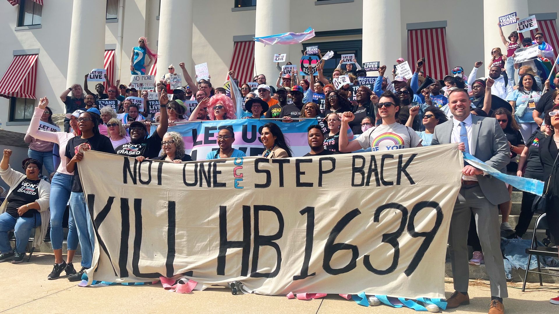 Several dozen people stand on the steps of a government building waving transgender pride flags and a banner that says "NOT ONE STEP BACK KILL HB 1639."