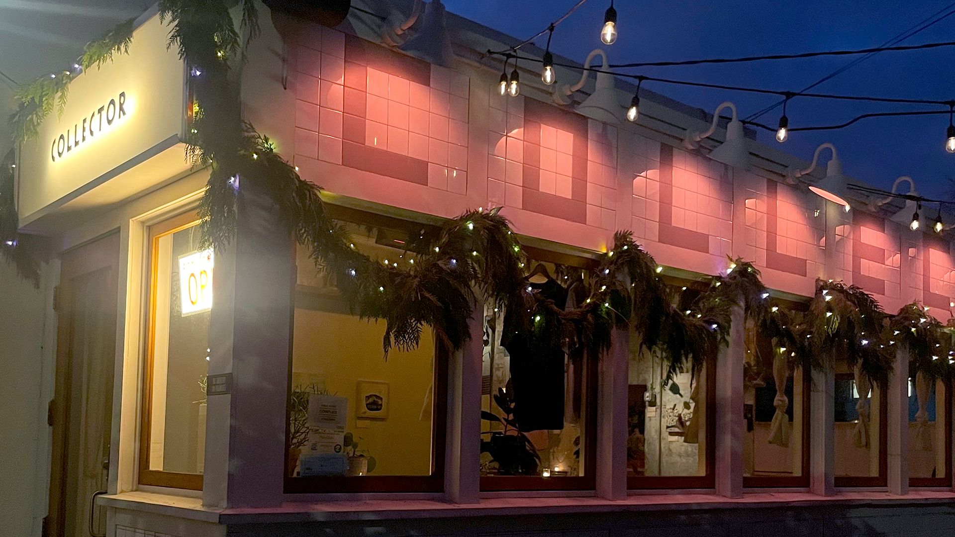 A cozy storefront at night with a sign reading "COLLECTOR," decorated with string lights and festive garlands. Warm light glows from inside, and pink tiles and hanging bulbs add charm to the exterior.  