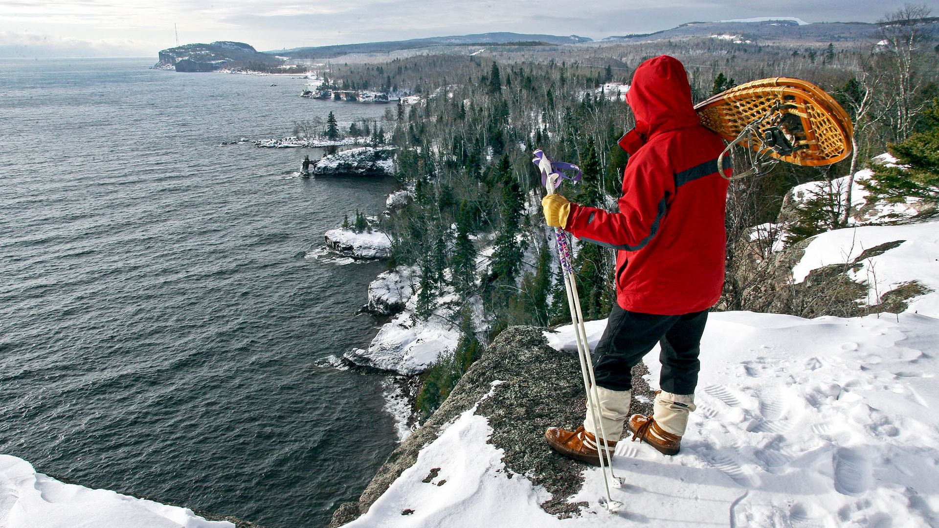 A snowshoer stands near a high rocky cliff overlooking the shore of Lake Superior with snow on the ground, green pines, and waves lapping under a cloudy sky