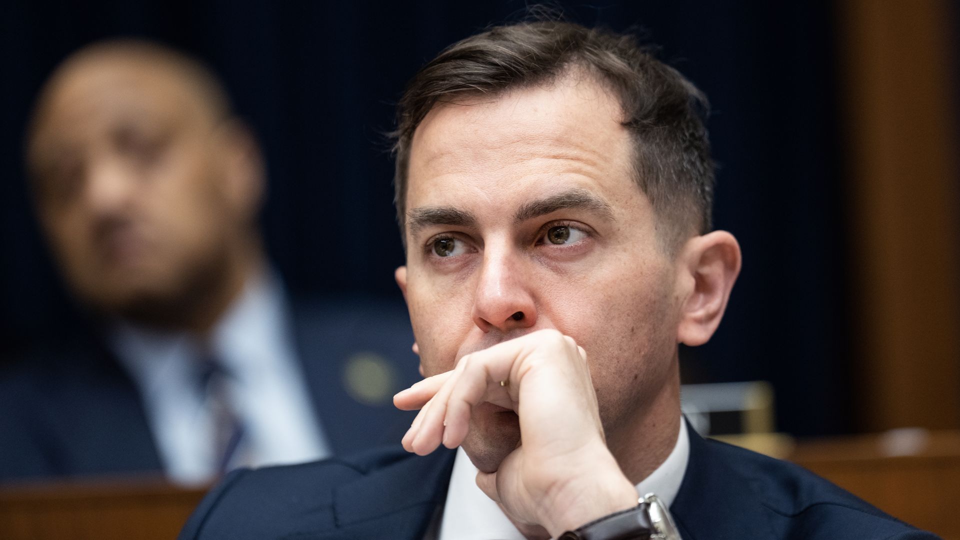 Rep. Rob Menendez, wearing a blue suit and holding his hand to his face while sitting at a brown wooden committee dais.