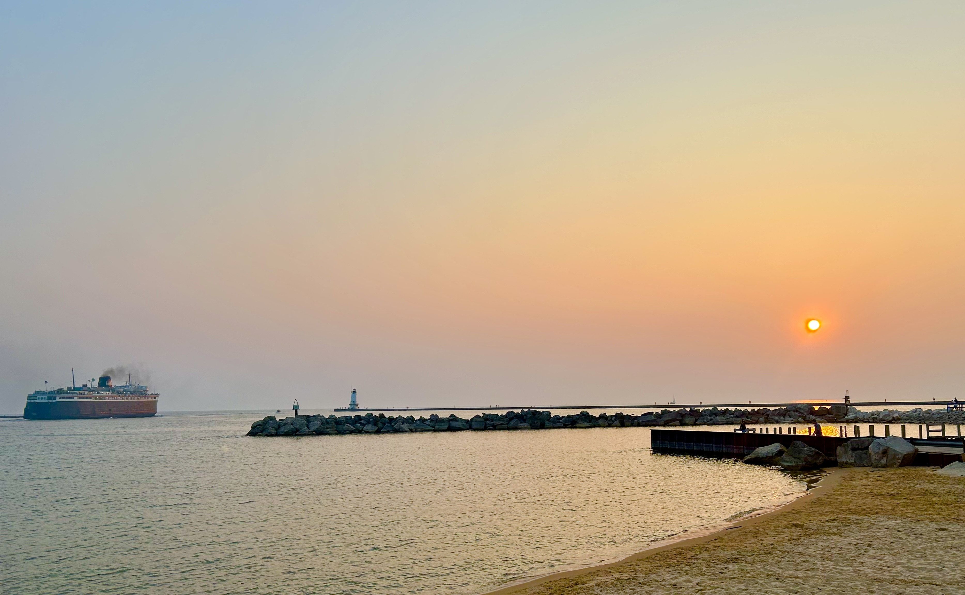 Orange sunset over calm water with a ship leaving a harbor, rocky breakwater, lighthouse, and people on a pier near sandy shore.