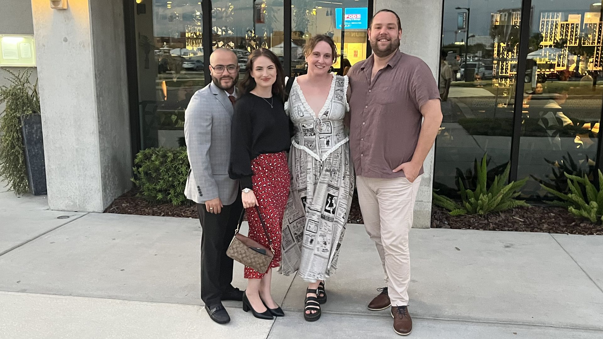 Four adults pose outside a glass storefront, smiling. From left: man in a gray blazer, woman in a black top with red patterned skirt, woman in a newspaper-print dress, and man in a mauve shirt with light pants.