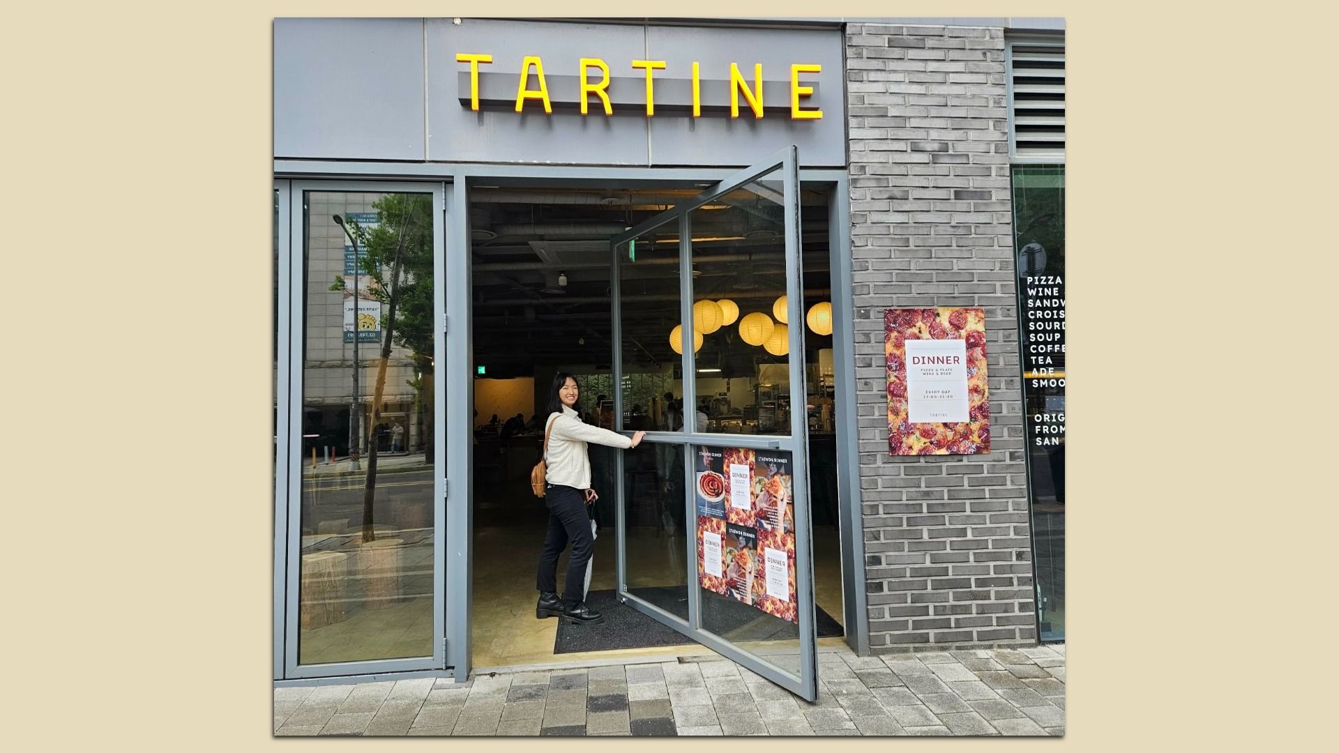 Front of a Tartine restaurant with bright yellow letters on a gray facade. A woman in a white jacket opens the glass door; brick wall to the right, and round warm lights inside.