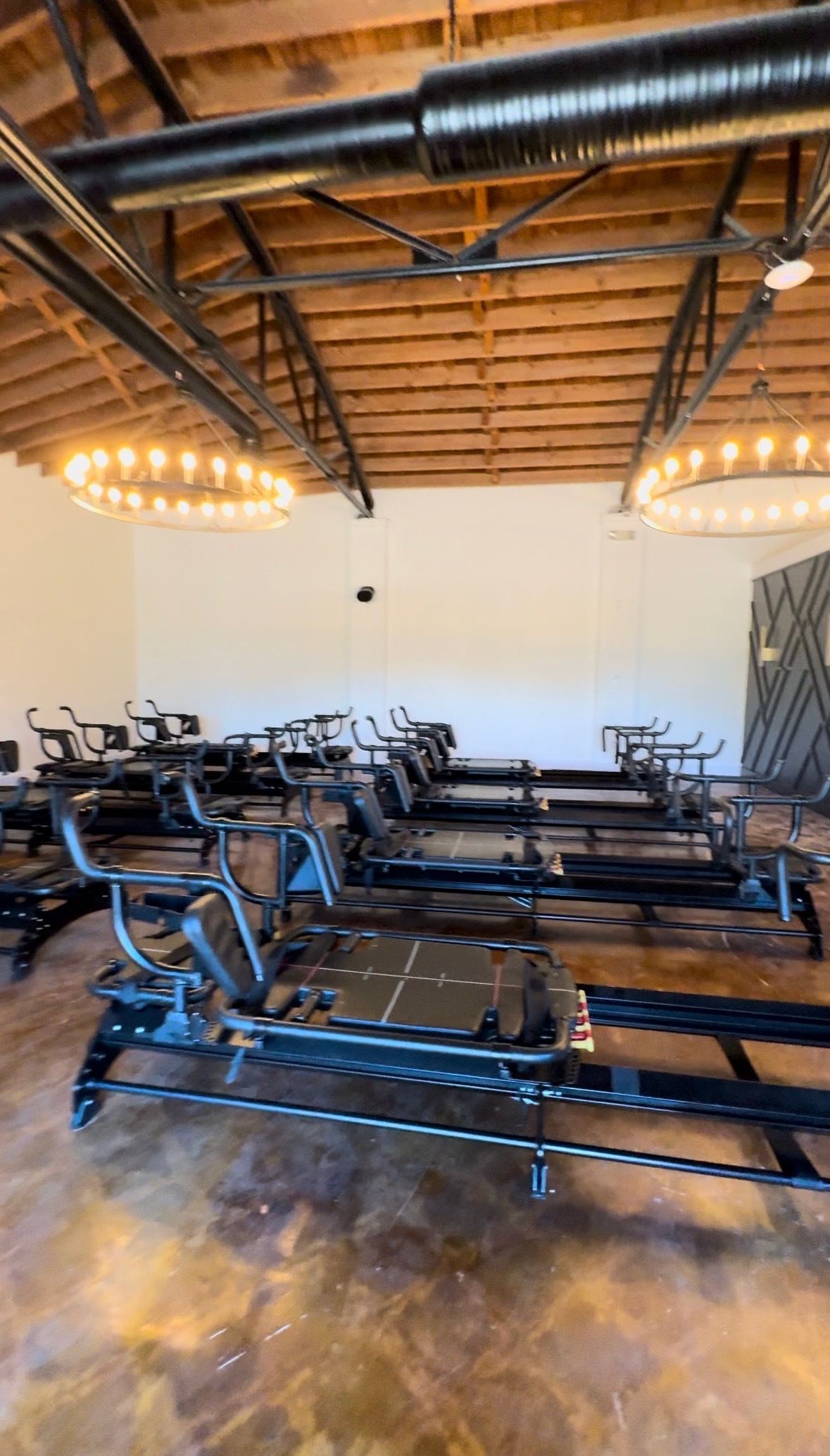 Room with multiple black Pilates reformer machines lined up on a polished brown floor, under wooden ceiling beams with black metal pipes and round chandeliers with warm lights.