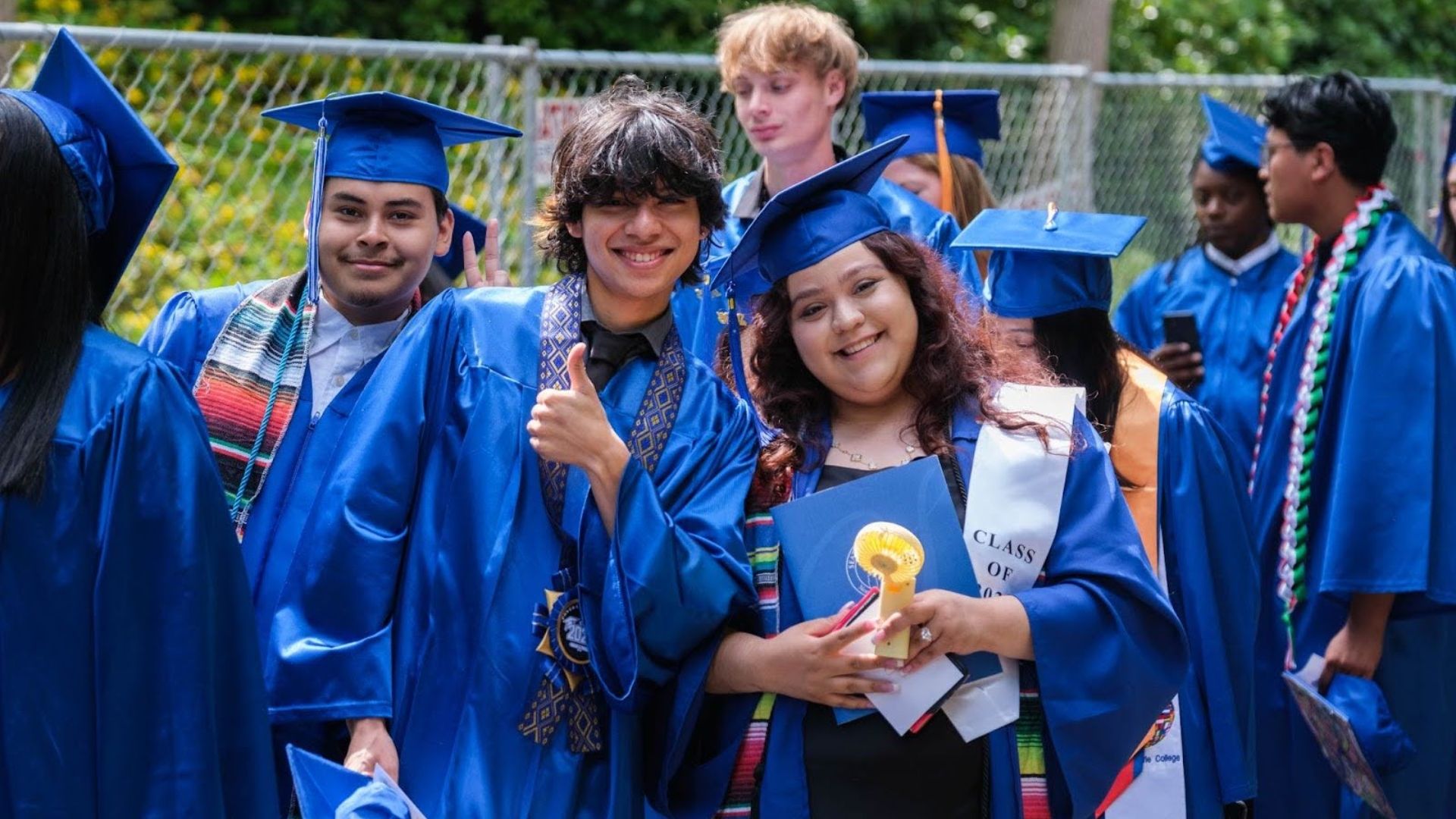 Group of graduates in blue gowns and caps celebrating outdoors, some wearing colorful stoles and sashes, smiling and giving thumbs up, with greenery and a fence in the background.